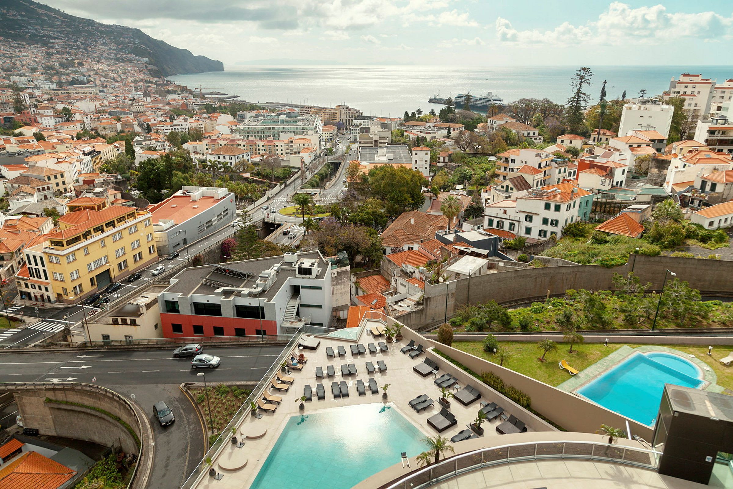 Udsigt over en by med bygninger, veje, to pools og havet i baggrunden. (AI-genereret tekst)