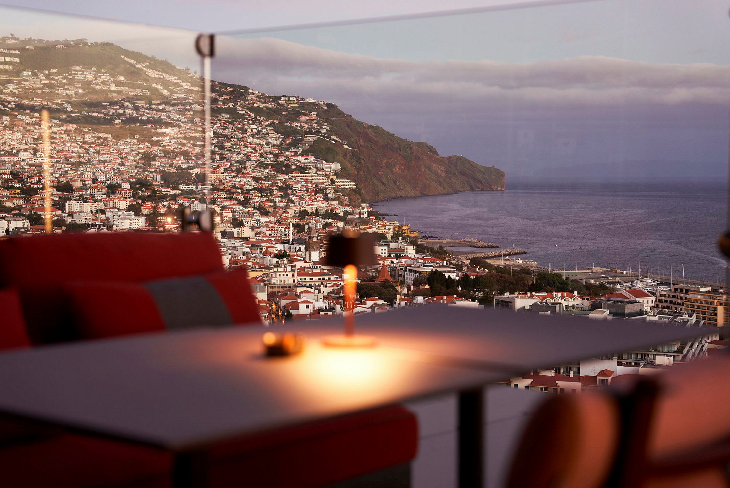 Udsigt over en kystby med en bjergskråning og havet i baggrunden, set fra en restaurant med røde siddepladser. (AI-genereret tekst)