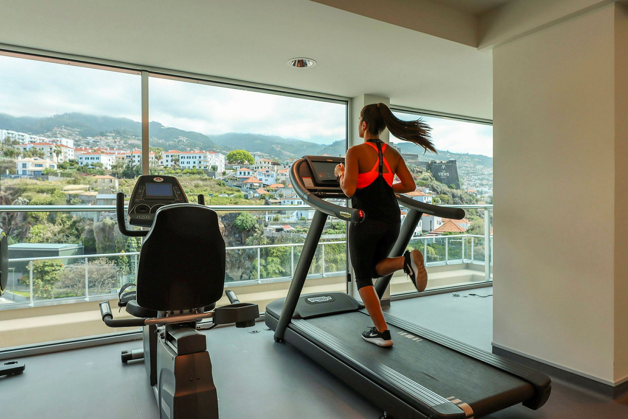 En person løber på løbebånd i et fitnesscenter med store vinduer og udsigt over byen. (AI-genereret tekst)