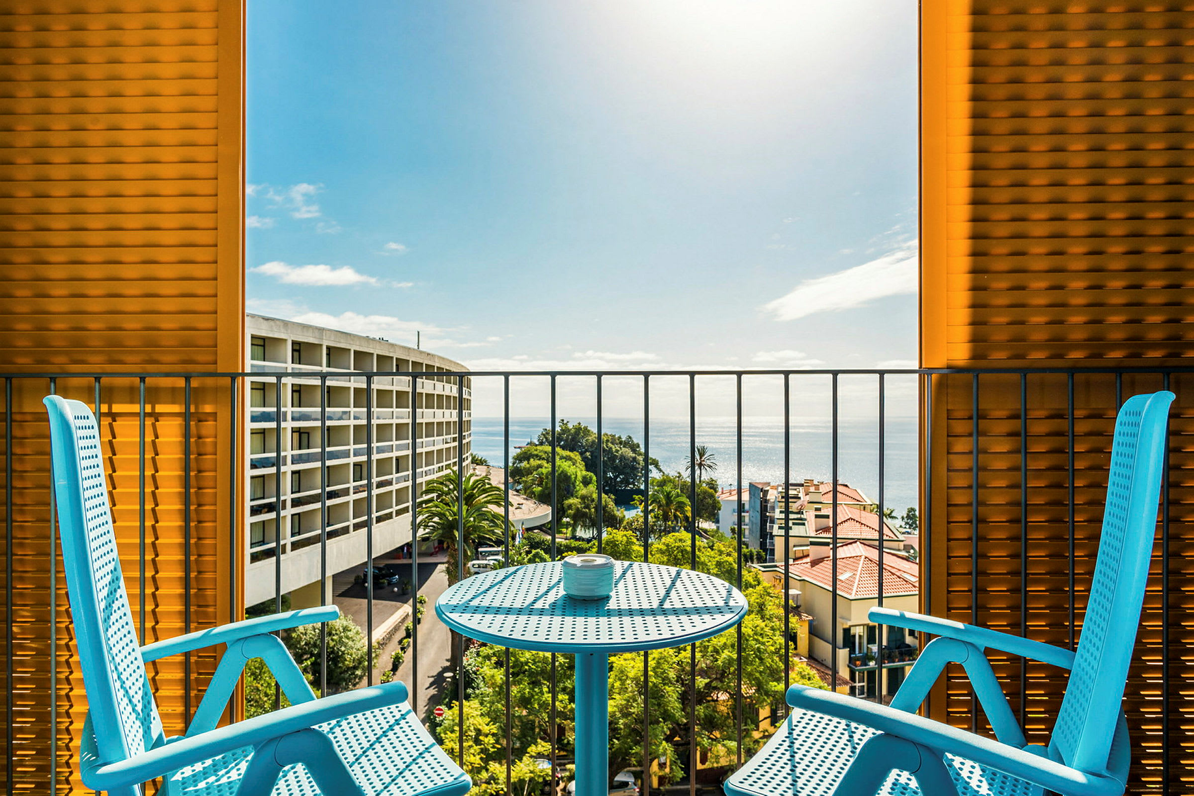 To blå stole og et bord på en balkon med udsigt over havet og omkringliggende bygninger. (AI-genereret tekst)