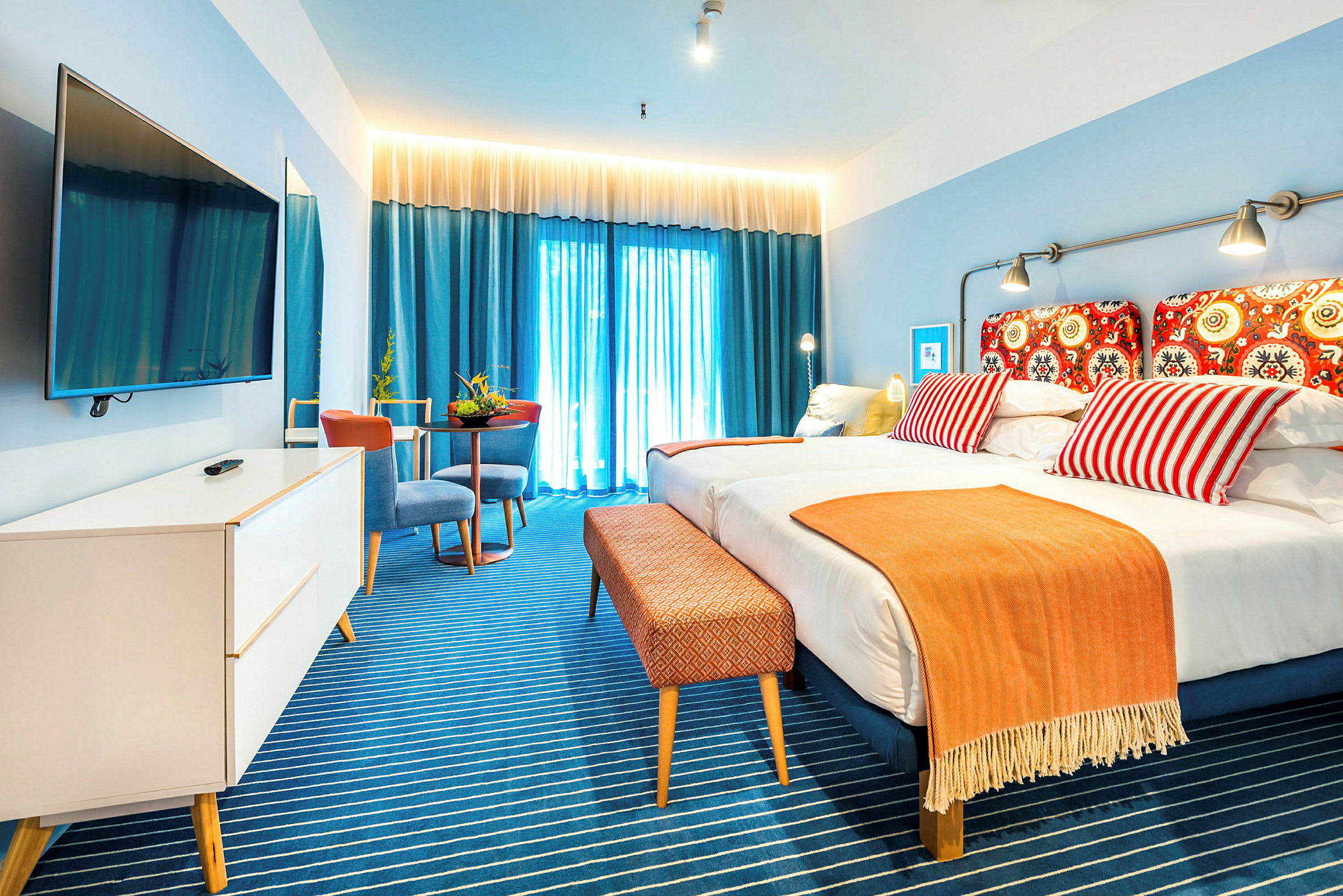 Hotelværelse med en dobbeltseng, farverige puder og et orange tæppe. Blå gardiner med lysindfald i baggrunden. (AI-genereret tekst)