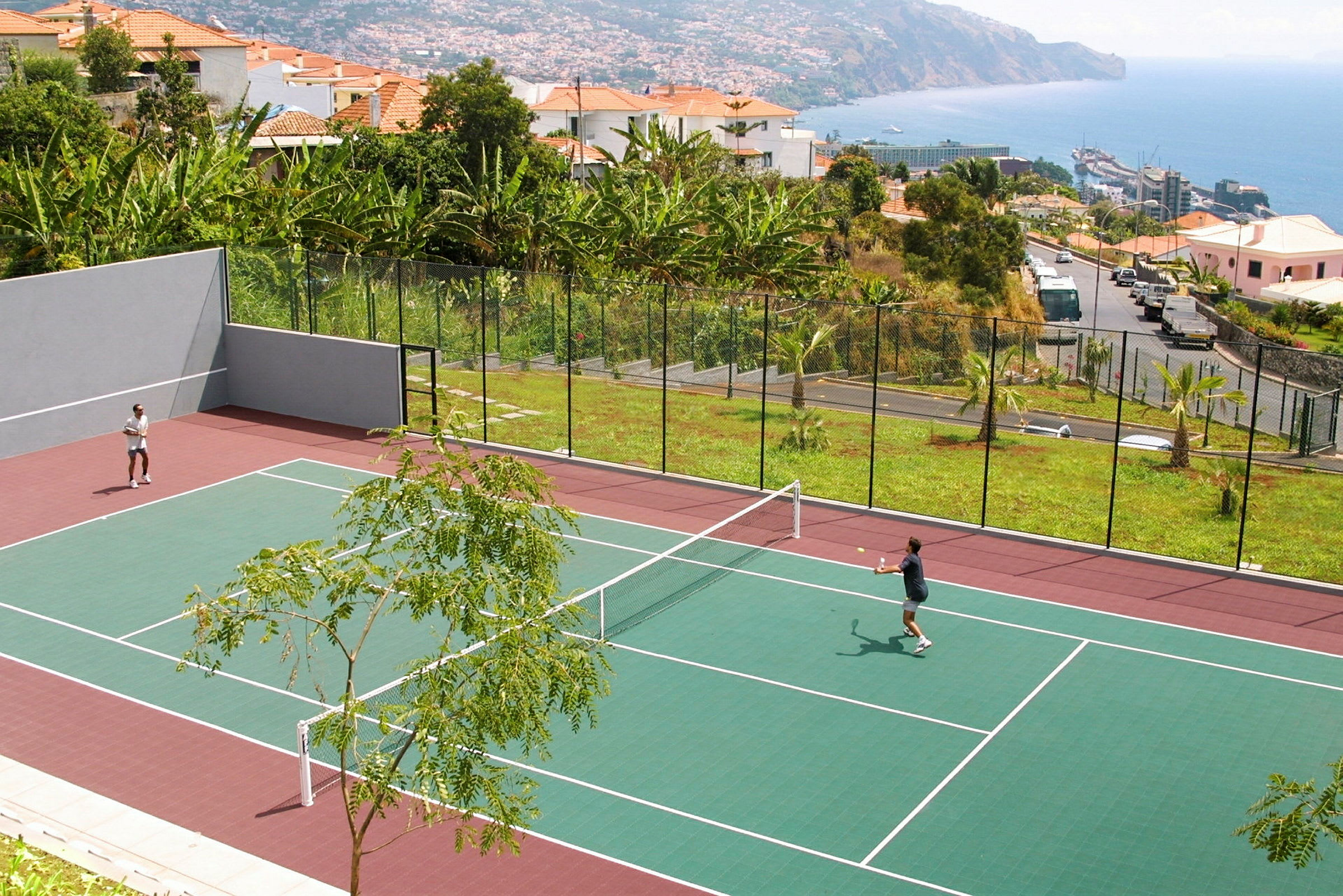 To personer spiller tennis på en udendørsbane med omkringliggende grønne områder og havudsigt. (AI-genereret tekst)