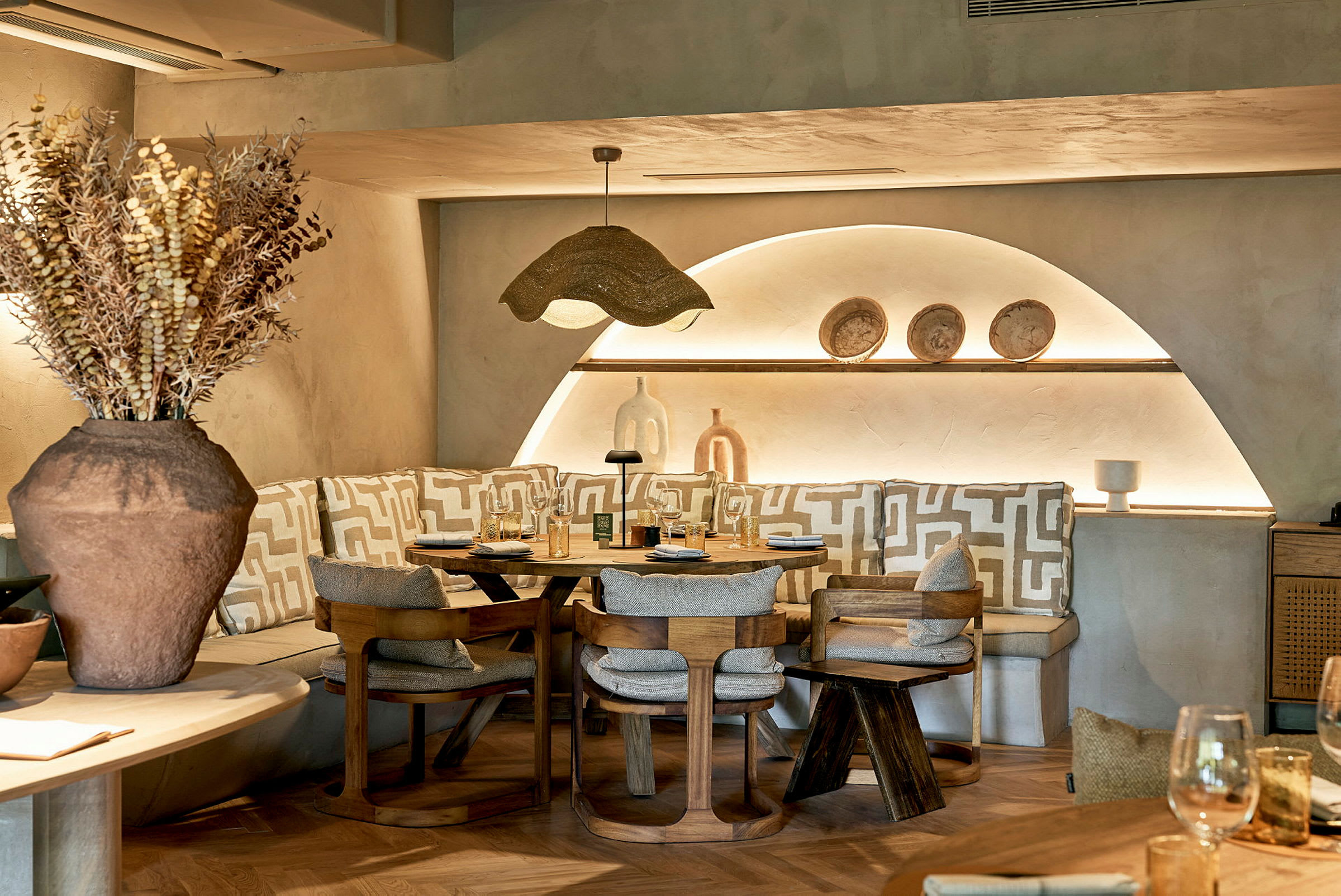 Et hyggeligt restaurantområde med en buet siddeplads, rustikke træstole og en dekorativ lampe over bordet. Keramiske pyntegenstande ses på hylden. (AI-genereret tekst)