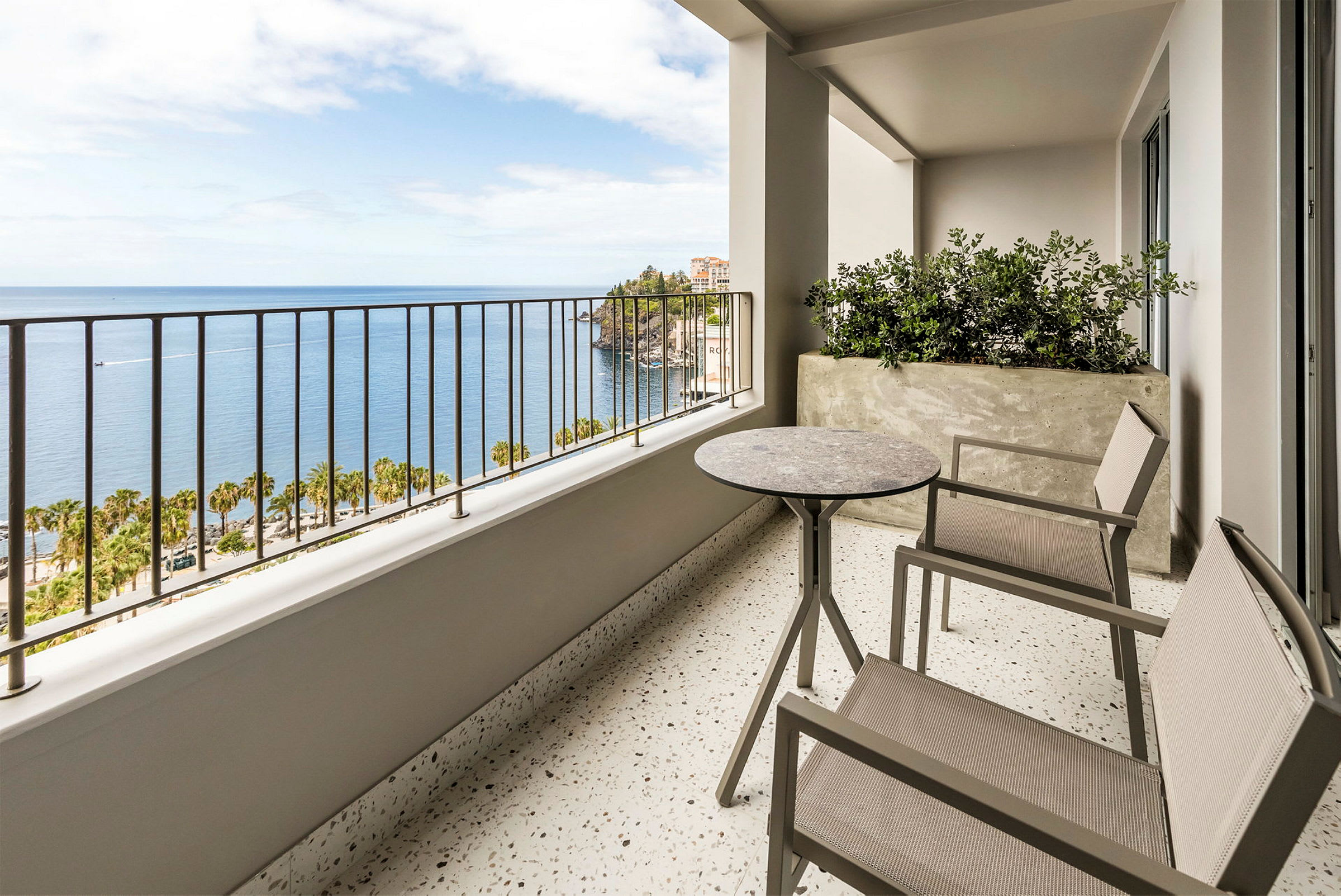 En balkon med to stole og et bord samt en udsigt over havet og palmer. (AI-genereret tekst)