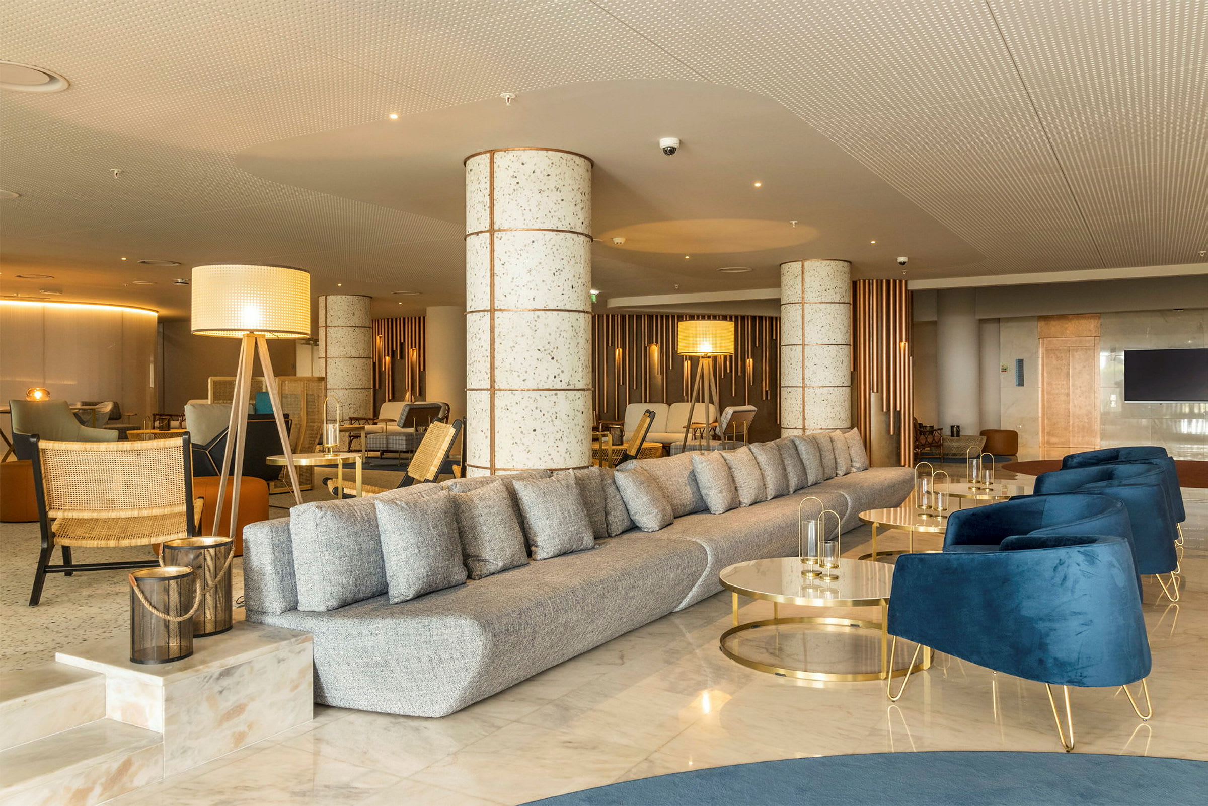 Et moderne indrettet hotellobbyområde med en lang grå sofa, blå lænestole og dekorative lysarmaturer. (AI-genereret tekst)