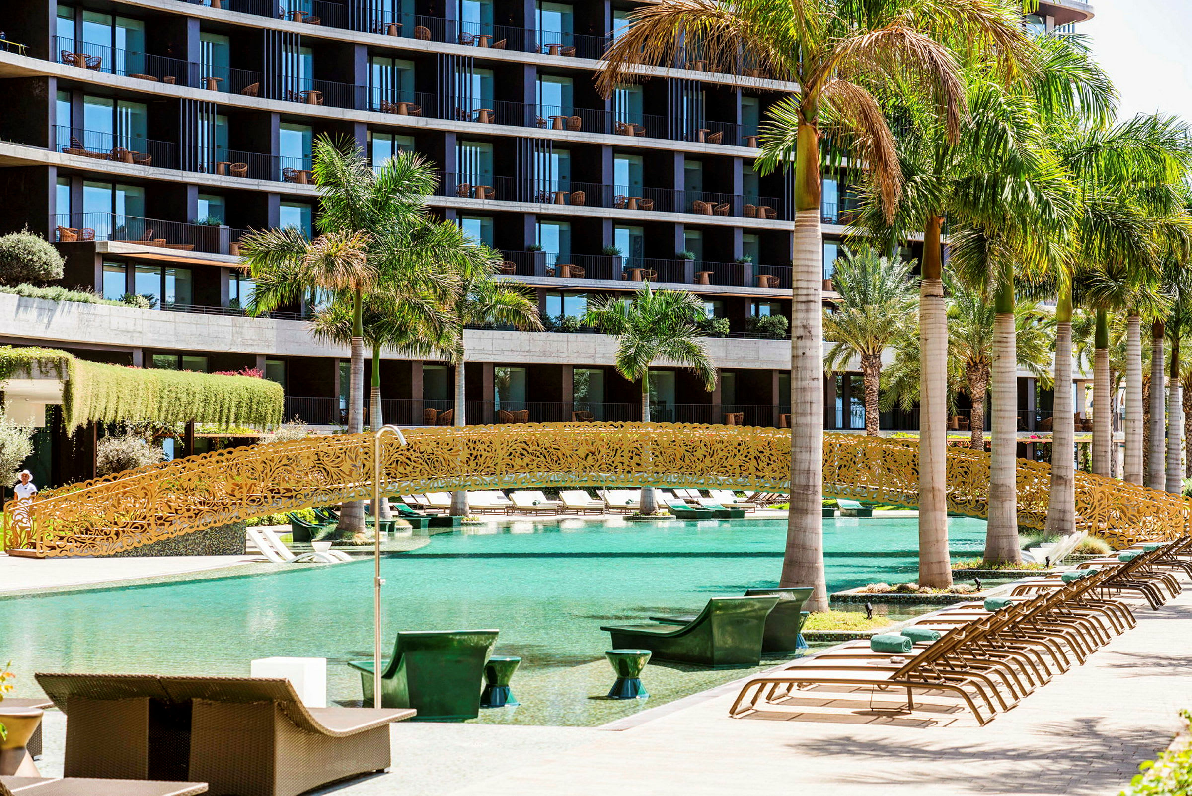 En pool med en dekorativ guldfarvet bro og omkringliggende palmer foran et moderne hotel. (AI-genereret tekst)