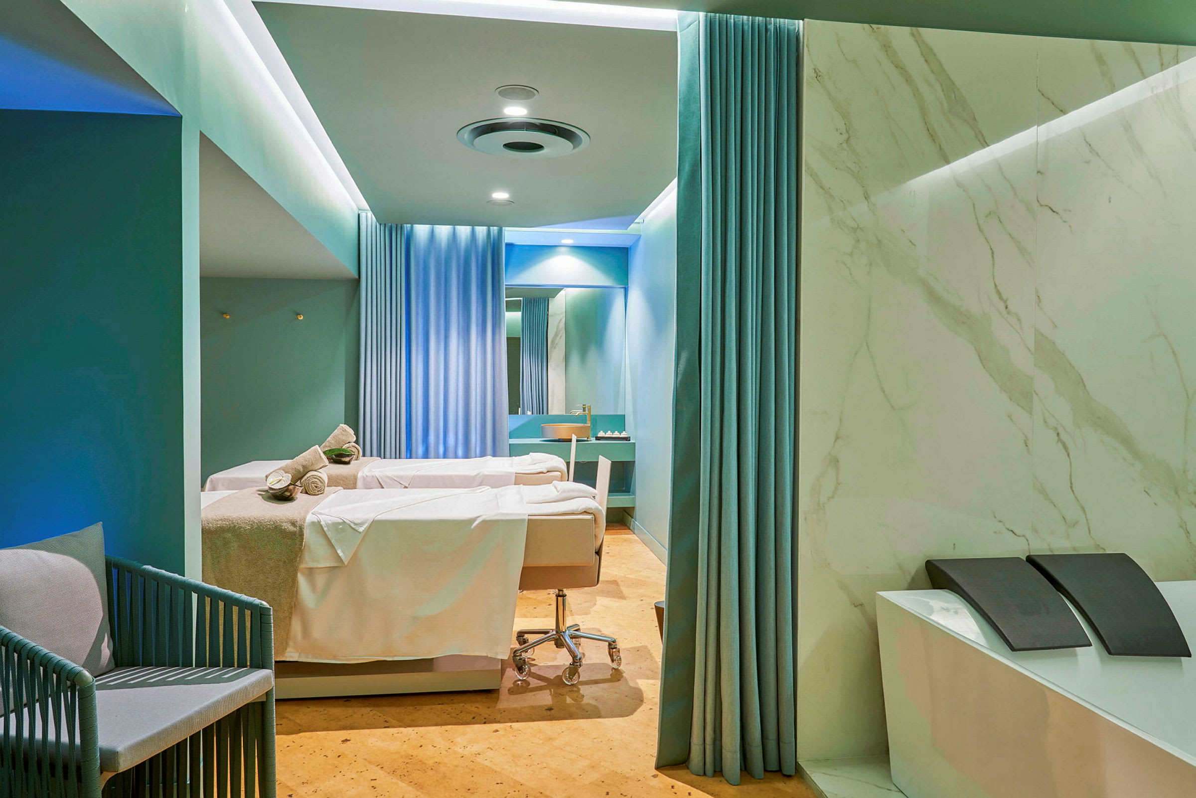 Et spa- eller behandlingsrum med to behandlingssenge dækket med hvide lagner og håndklæder. Rummet er indrettet med blå gardiner, marmorerede vægge og har et moderne design. (AI-genereret tekst)
