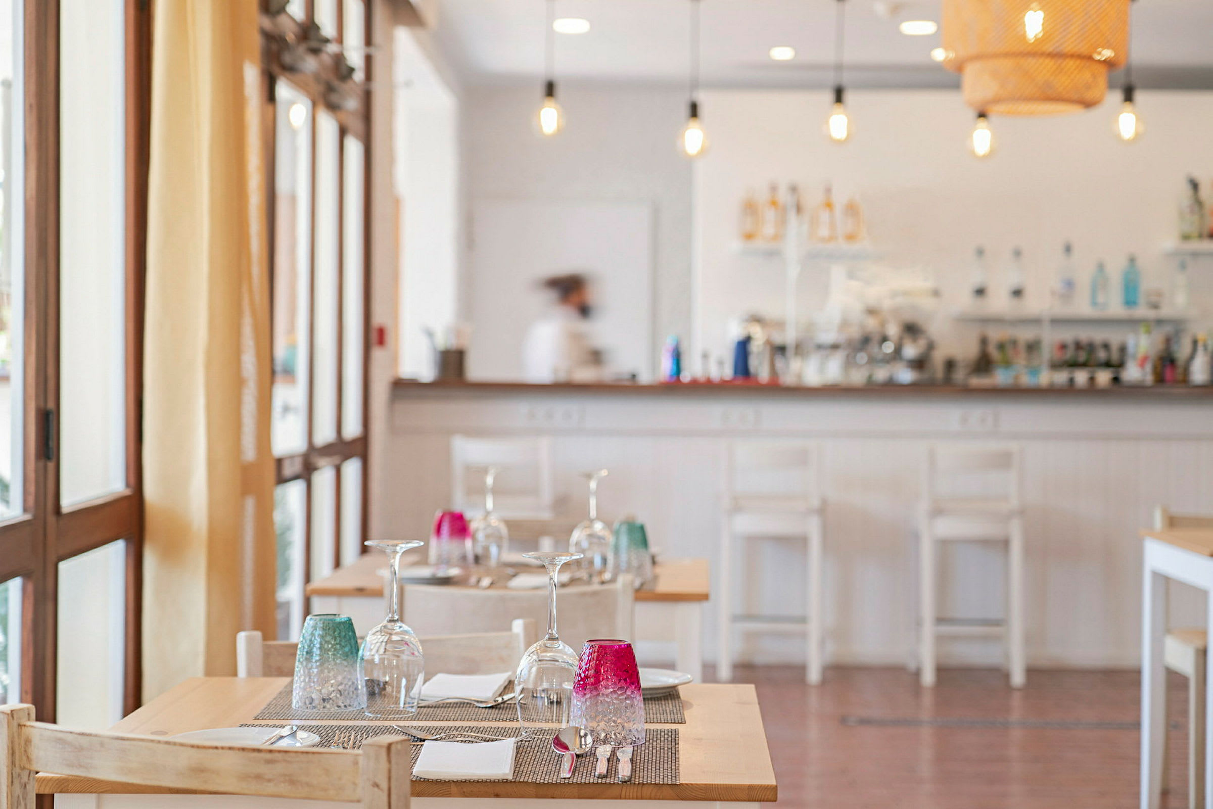 Interiør af en restaurant med dækkede borde med glas og farverige glas på. Tjener i baggrunden ved bardisken. (AI-genereret tekst)