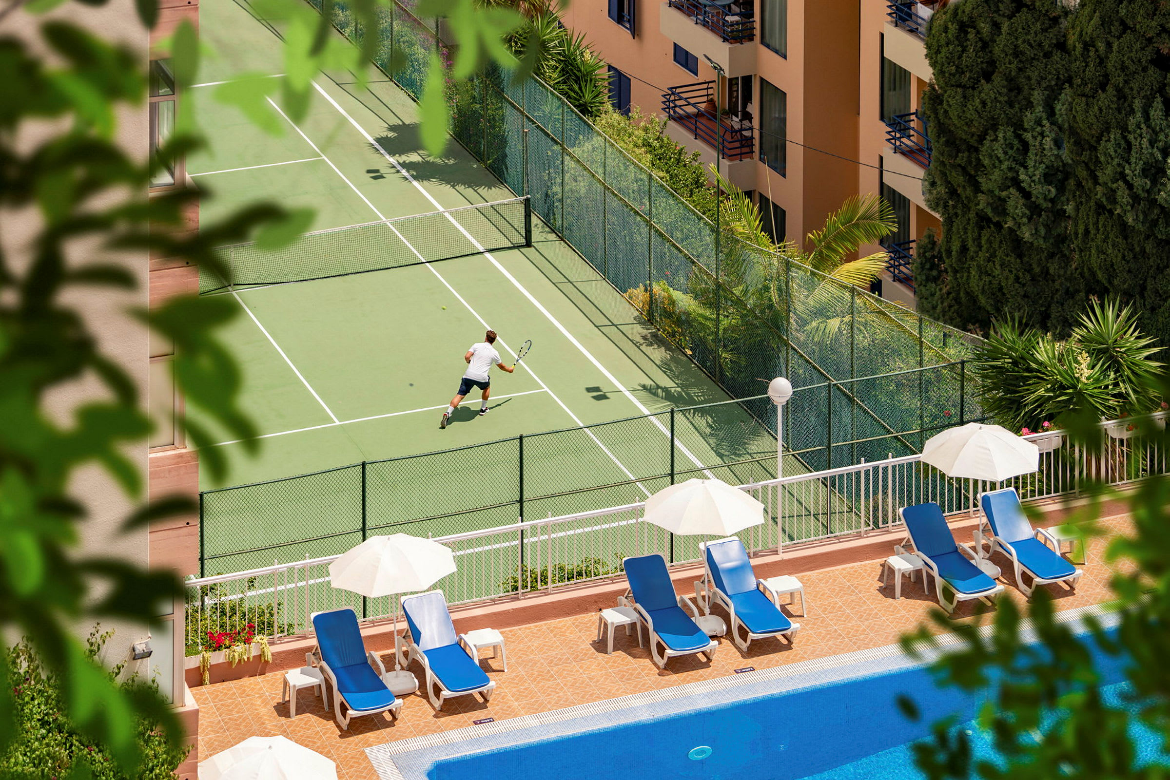 En person spiller tennis på en tennisbane tæt på en pool med liggestole og parasoller. (AI-genereret tekst)