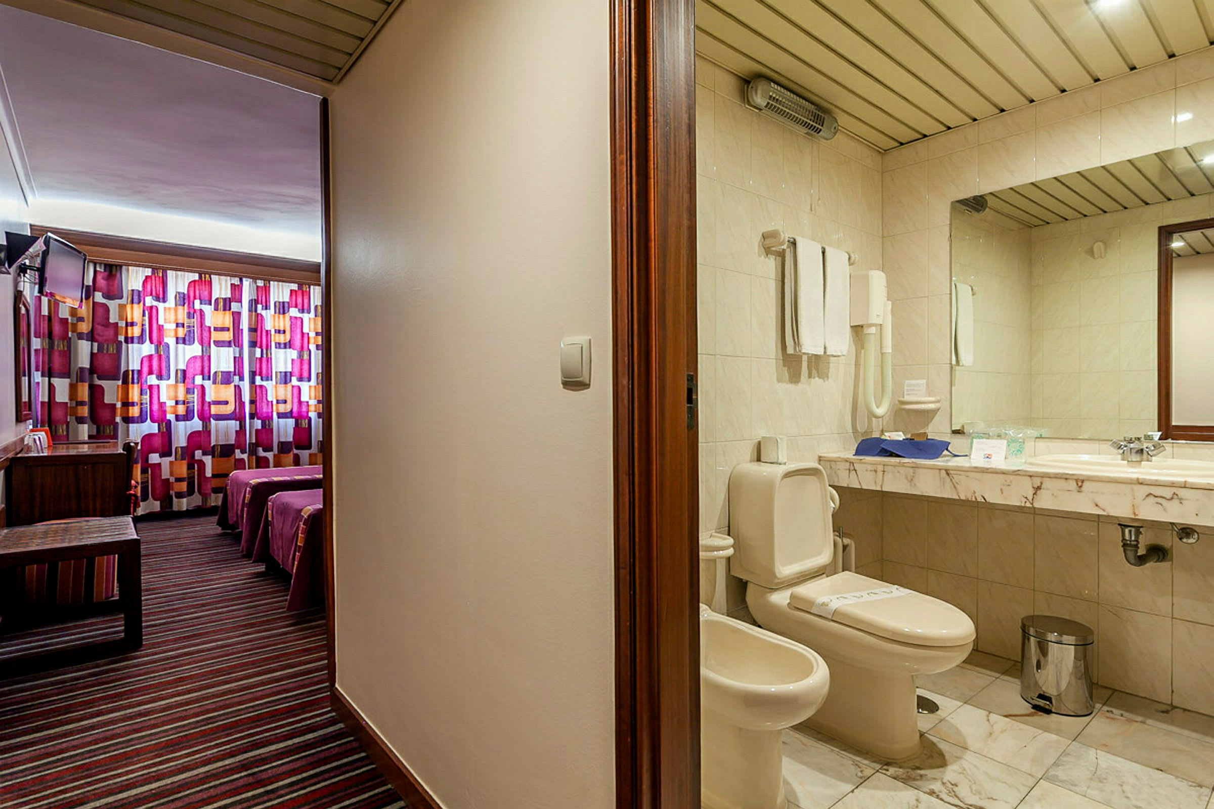 Et hotelværelsesinteriør, der viser et badeværelse med toilet og vask til venstre og et soveværelse med farverige gardiner og to senge til højre. (AI-genereret tekst)