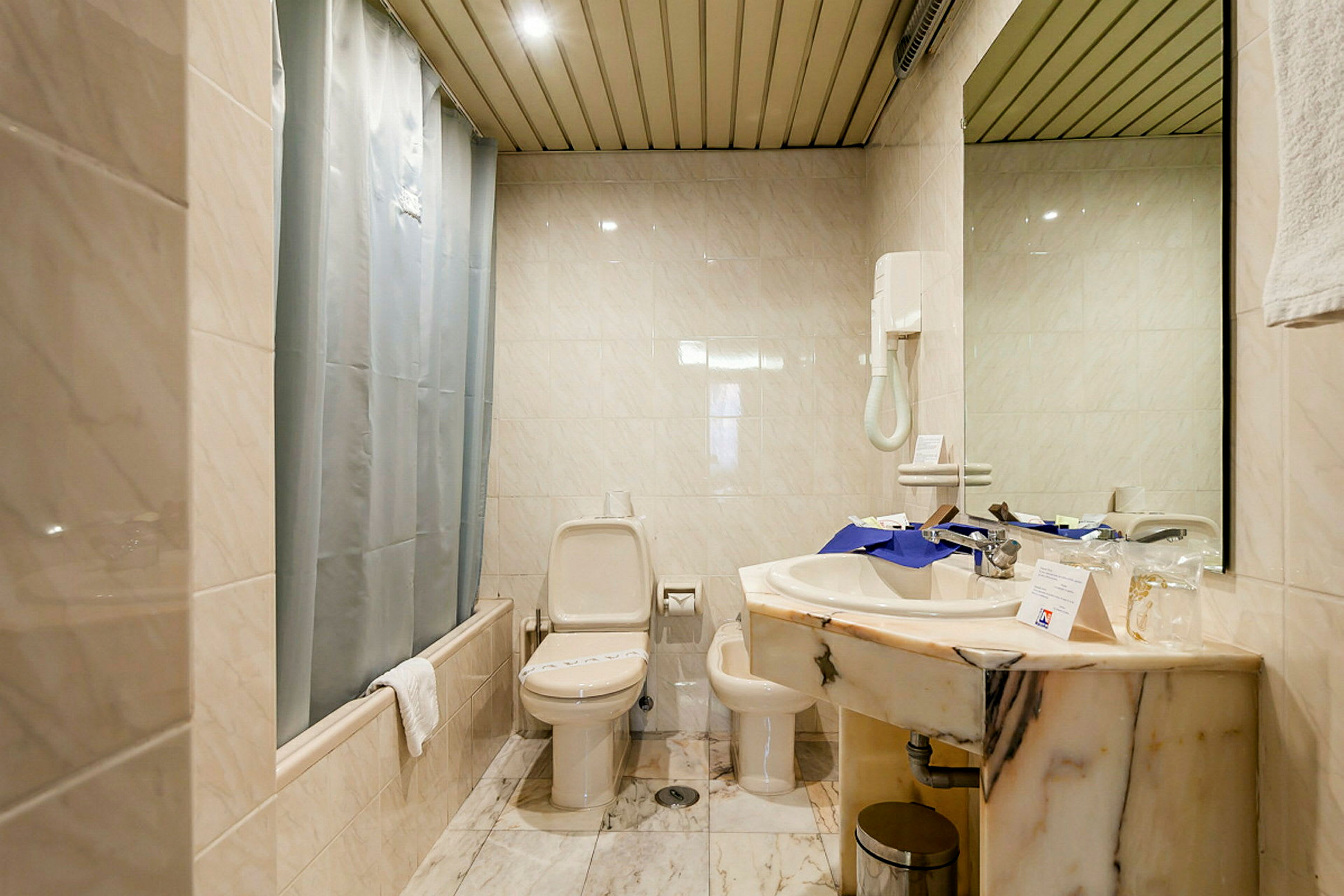 Et badeværelse med toilet, bidet og håndvask. Fliser på vægge og gulv samt en bruser med forhæng. (AI-genereret tekst)