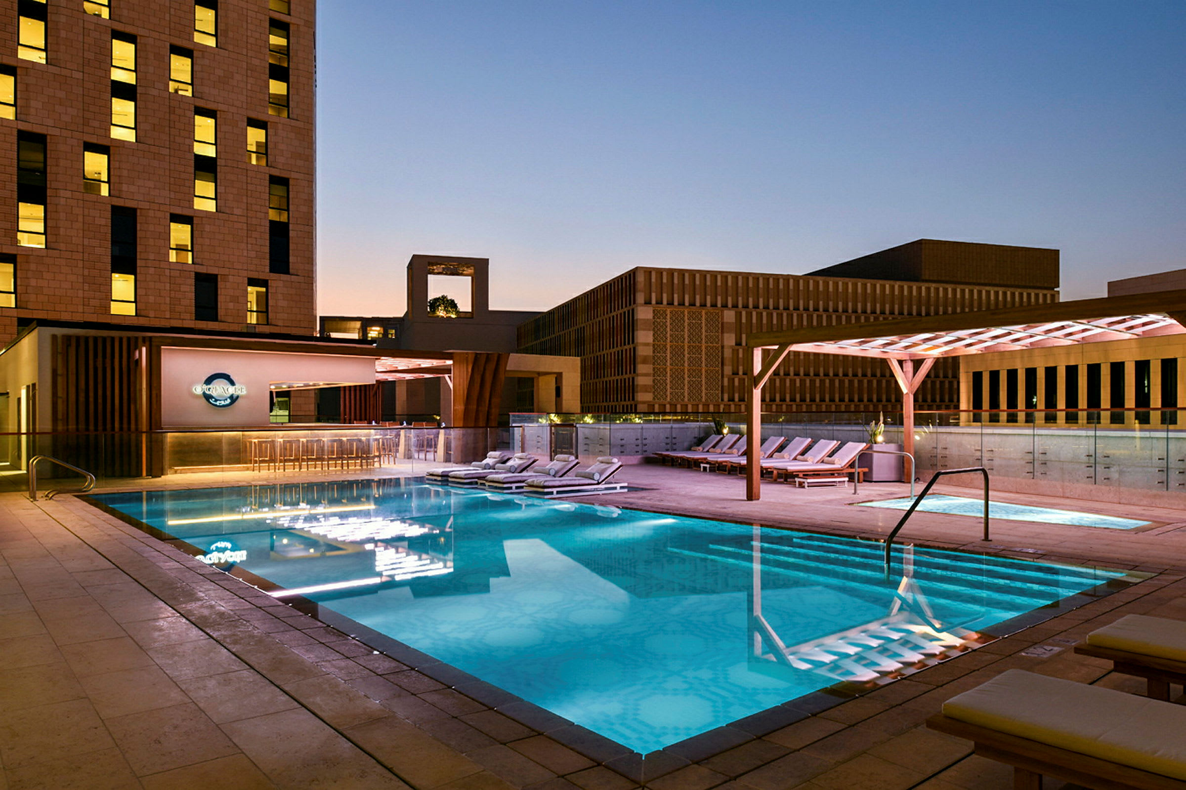 En oplyst udendørs swimmingpool om aftenen omgivet af liggestole og moderne bygningsarkitektur. (AI-genereret tekst)