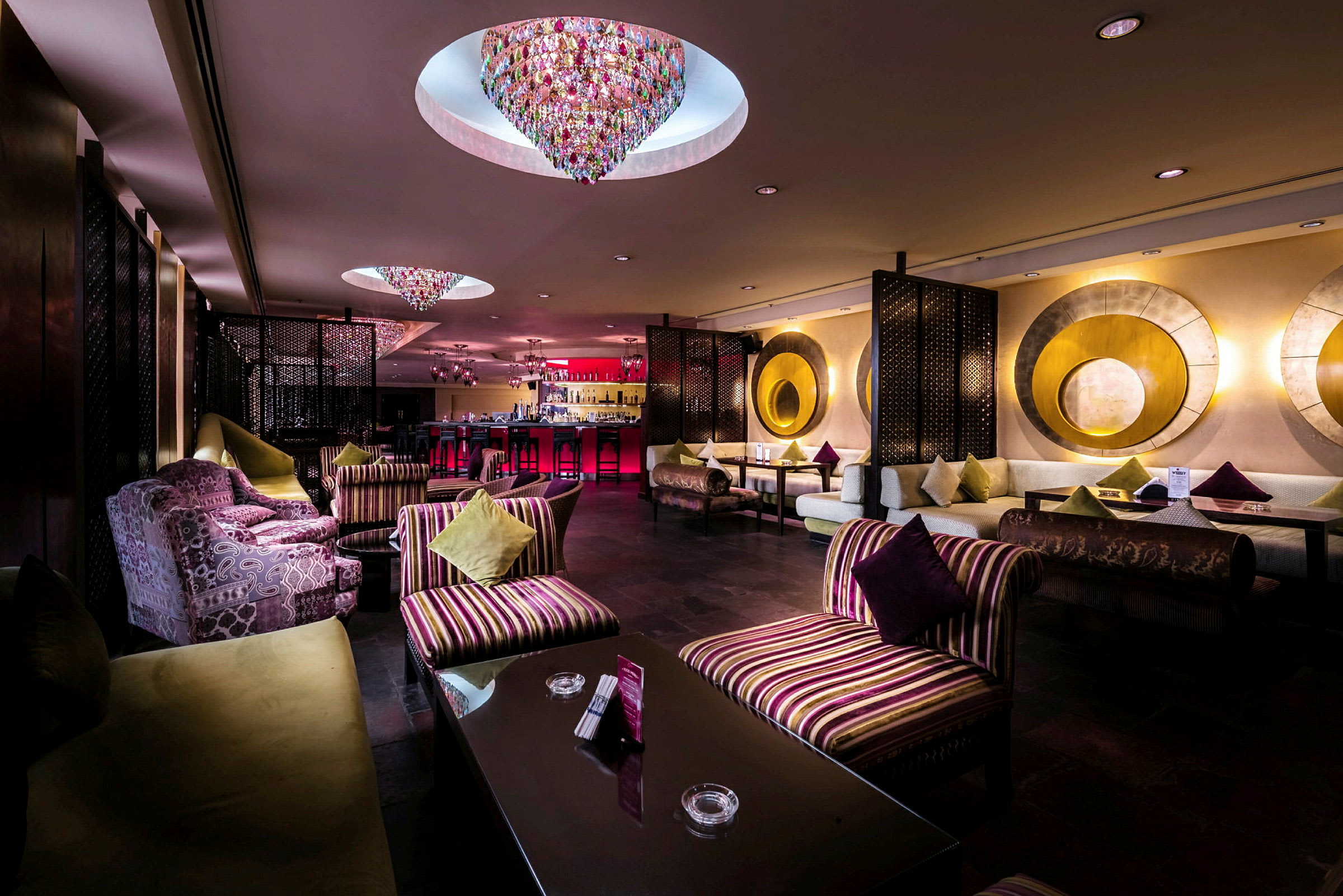 En elegant hotelbar med farverige lysekroner i loftet, bløde siddepladser og sofaer prydet med dekorative puder. (AI-genereret tekst)