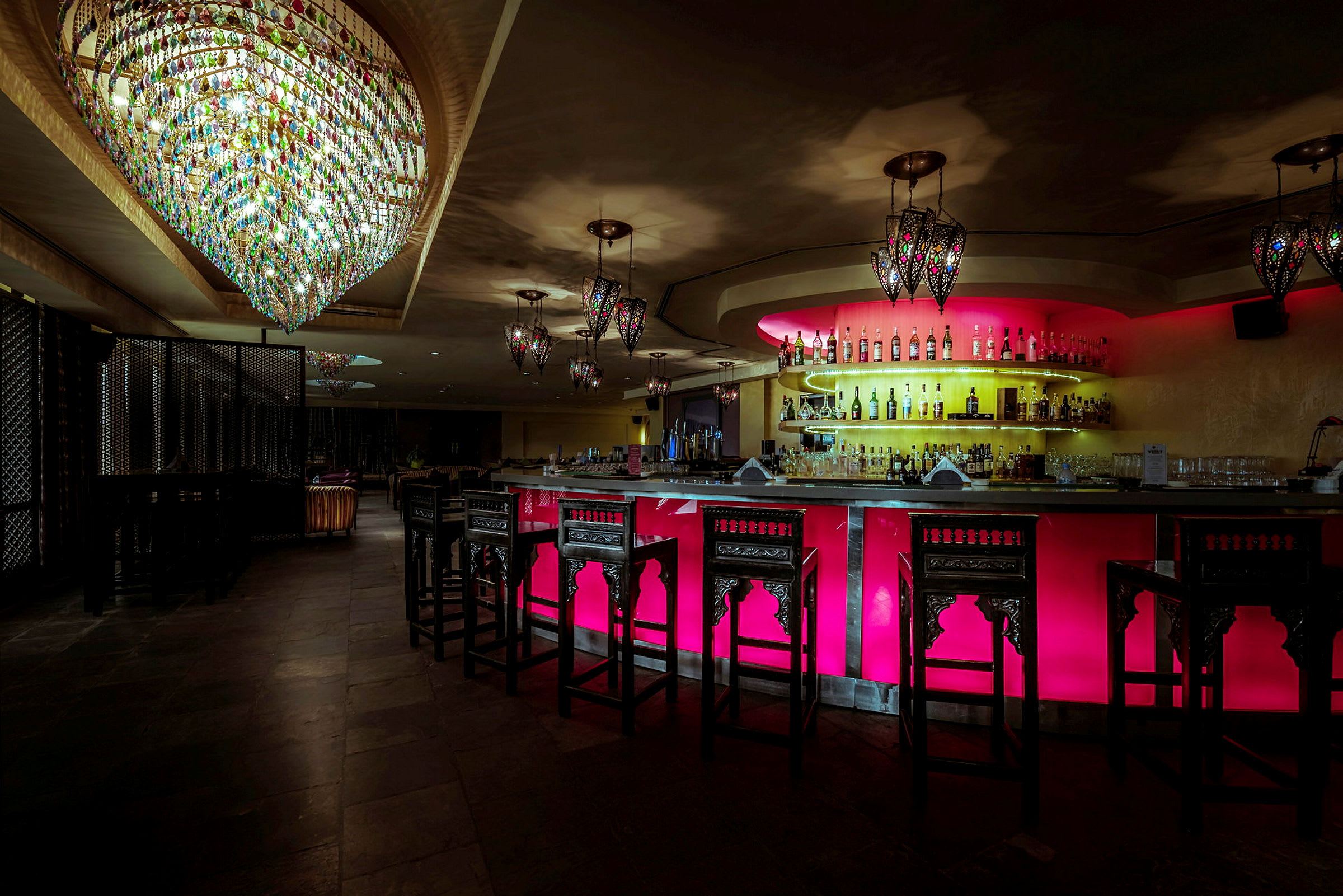 En elegant bar med oplyst bardisk i farverige toner og lysekroner hængende fra loftet. (AI-genereret tekst)