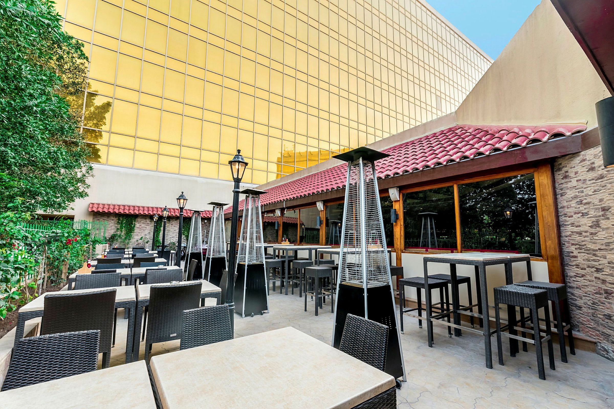 En udendørs terrasse med borde og stole, omgivet af grønne planter og en bygning med gylden facade. (AI-genereret tekst)