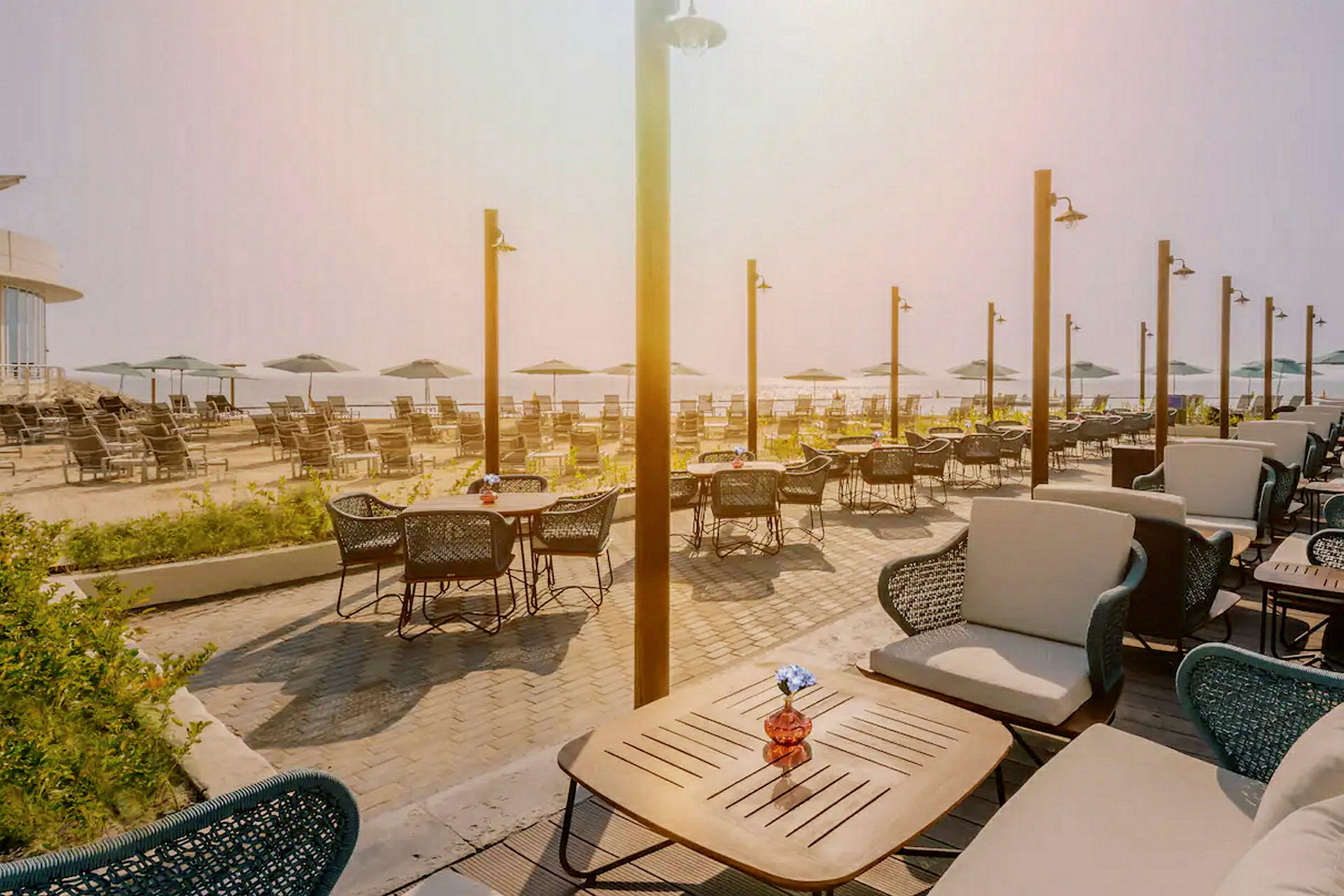 En udendørs café med borde og stole ved siden af parasoller og liggestole på en strand. (AI-genereret tekst)