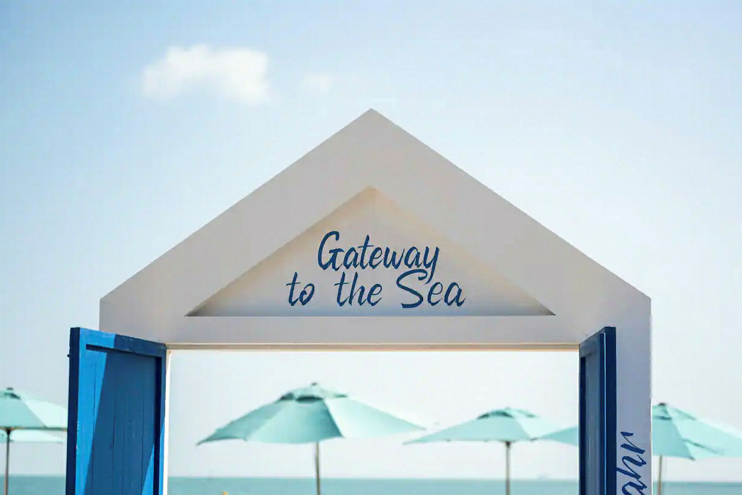 En hvid portal med teksten "Gateway to the Sea" med udsigt over havet og parasoller i baggrunden. (AI-genereret tekst)