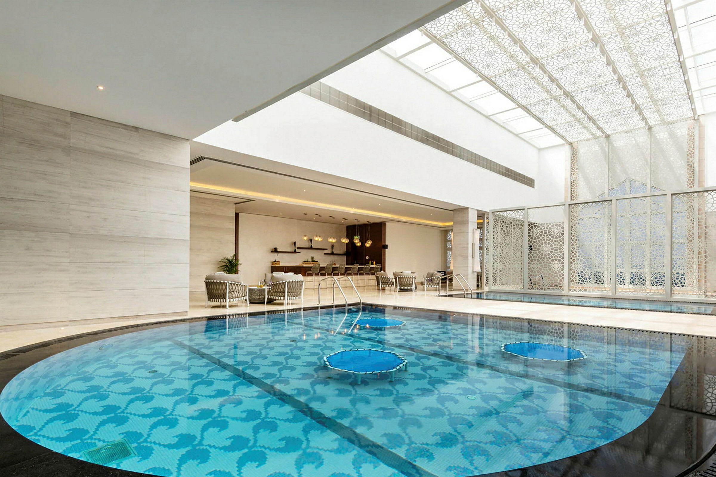 En indendørs pool med et dekorativt mønster på bunden, omgivet af en elegant lounge med siddepladser og et loft med stilfuldt designede paneler. (AI-genereret tekst)