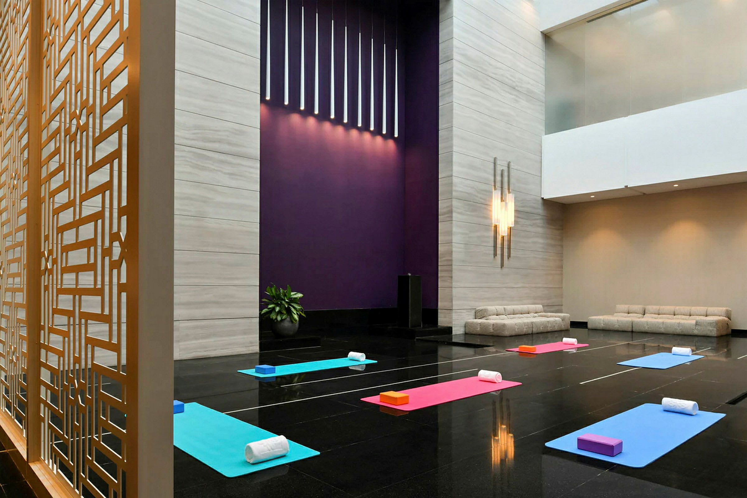 Et rum med yogamåtter og tilhørende tilbehør opstillet på et blankt gulv. Der er moderne sofaer til siden og en dekorativ gitteradskillelse. (AI-genereret tekst)