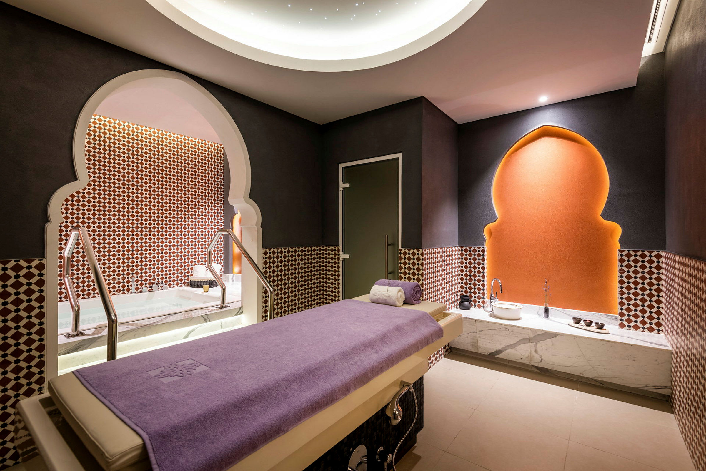 Et spa-rum med en massagebriks dækket af et lilla håndklæde, dekorative mønstre på væggene og en oplyst bue bagved. (AI-genereret tekst)
