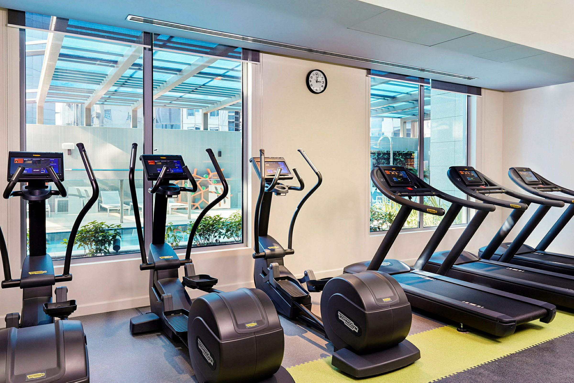 Et fitnessområde med løbebånd og træningsmaskiner ved siden af store vinduer. (AI-genereret tekst)