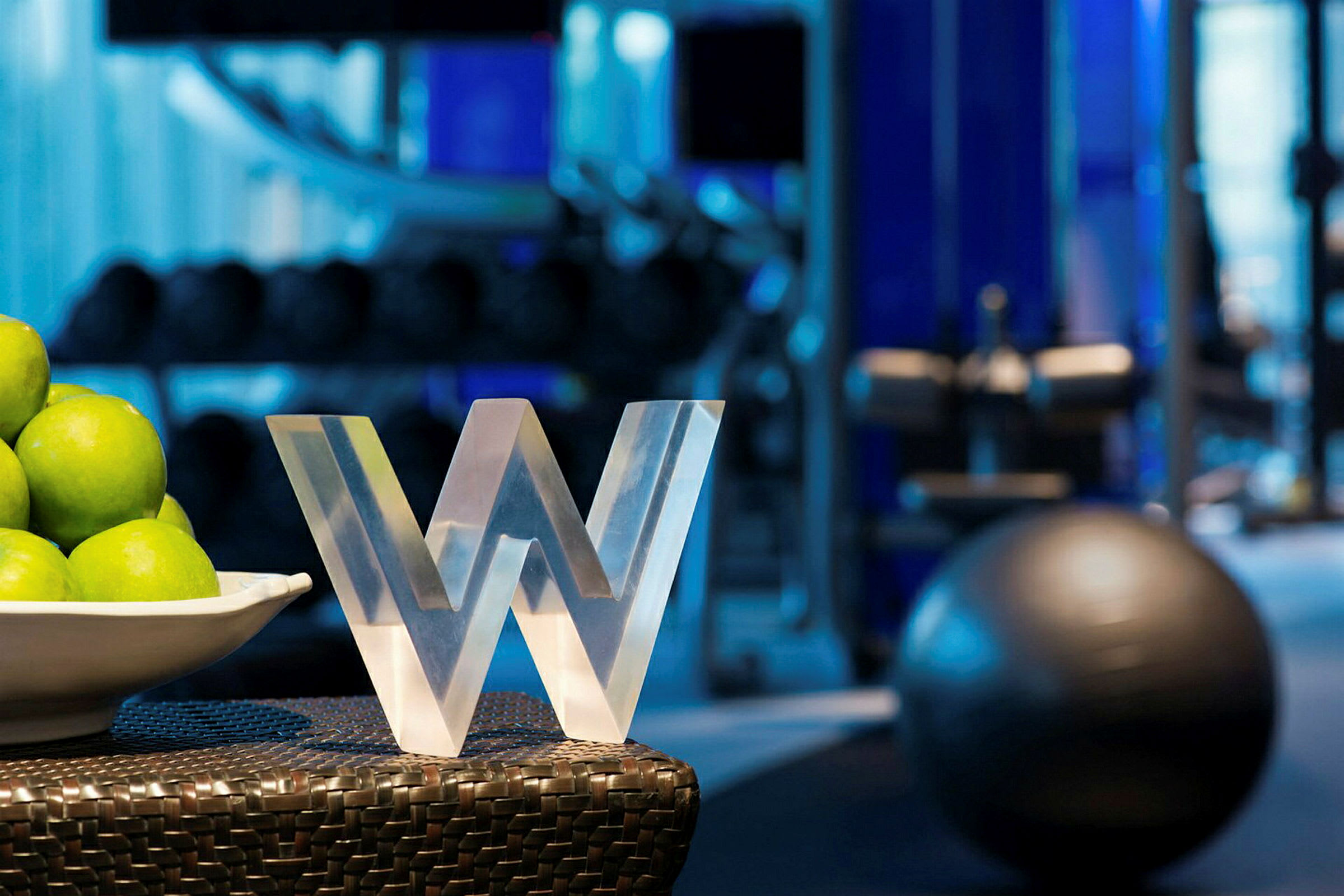 Et nærbillede af en skulptur formet som bogstavet 'W', der står ved siden af en hvid skål fyldt med grønne æbler. I baggrunden ses træningsudstyr i et fitnesscenter. (AI-genereret tekst)