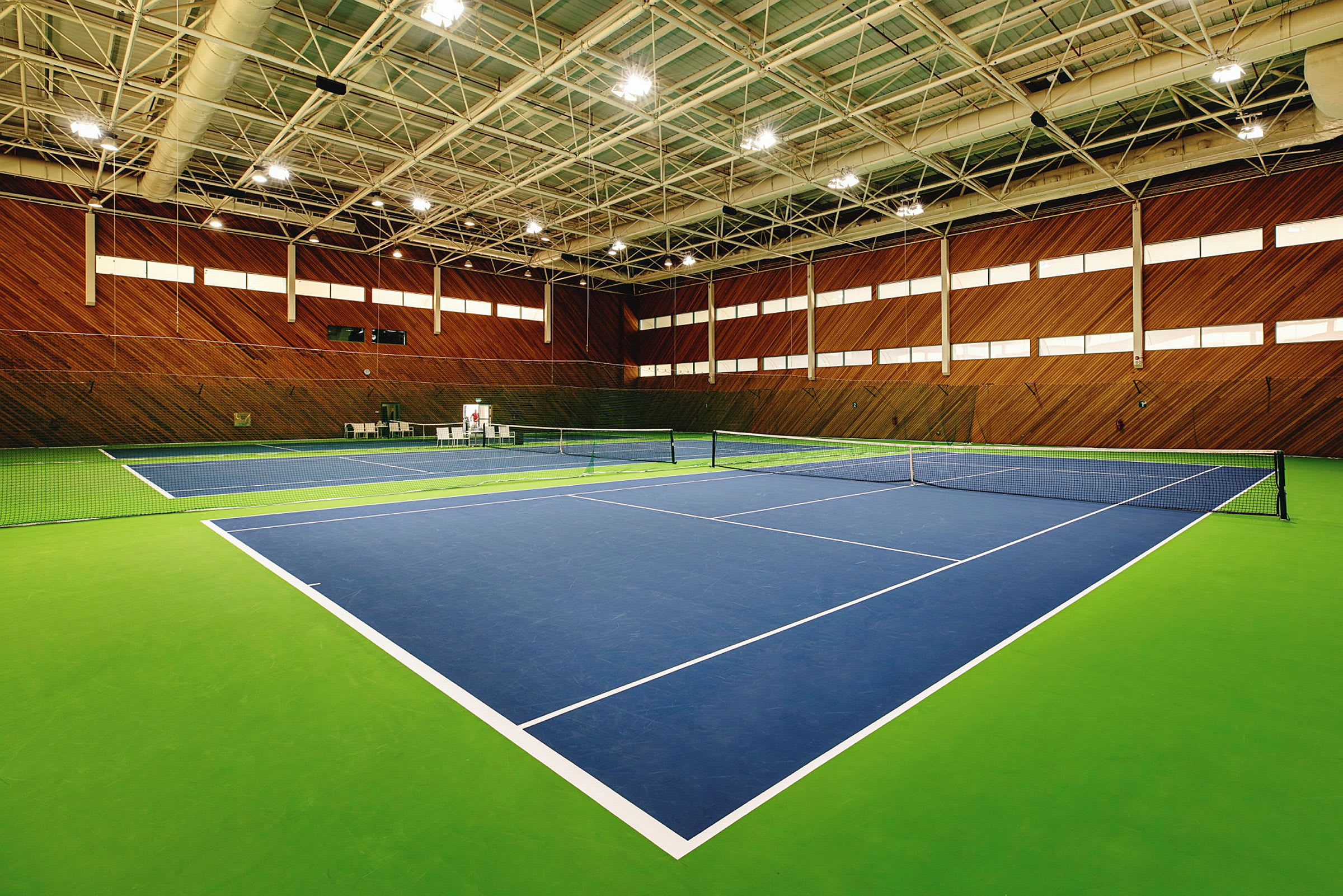 Et indendørs tennishal med to tennisbaner, malet i blåt og grønt, omgivet af træpaneler på væggene. (AI-genereret tekst)