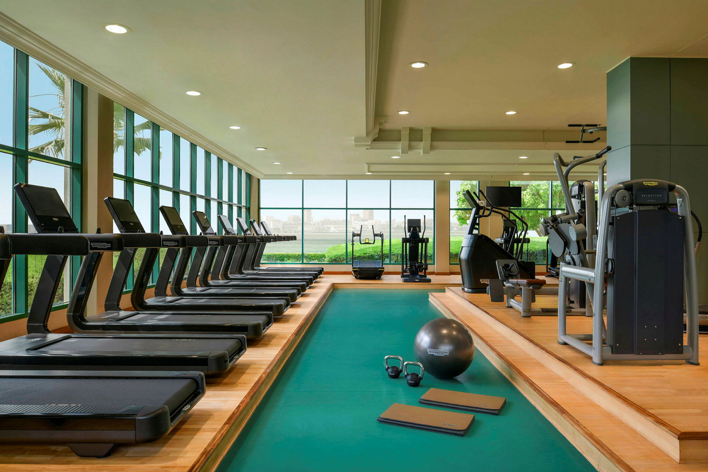 Et fitnesscenter med løbebånd, styrketræningsmaskiner og træningsudstyr. Store vinduer giver udsyn til grønne områder og byen. (AI-genereret tekst)