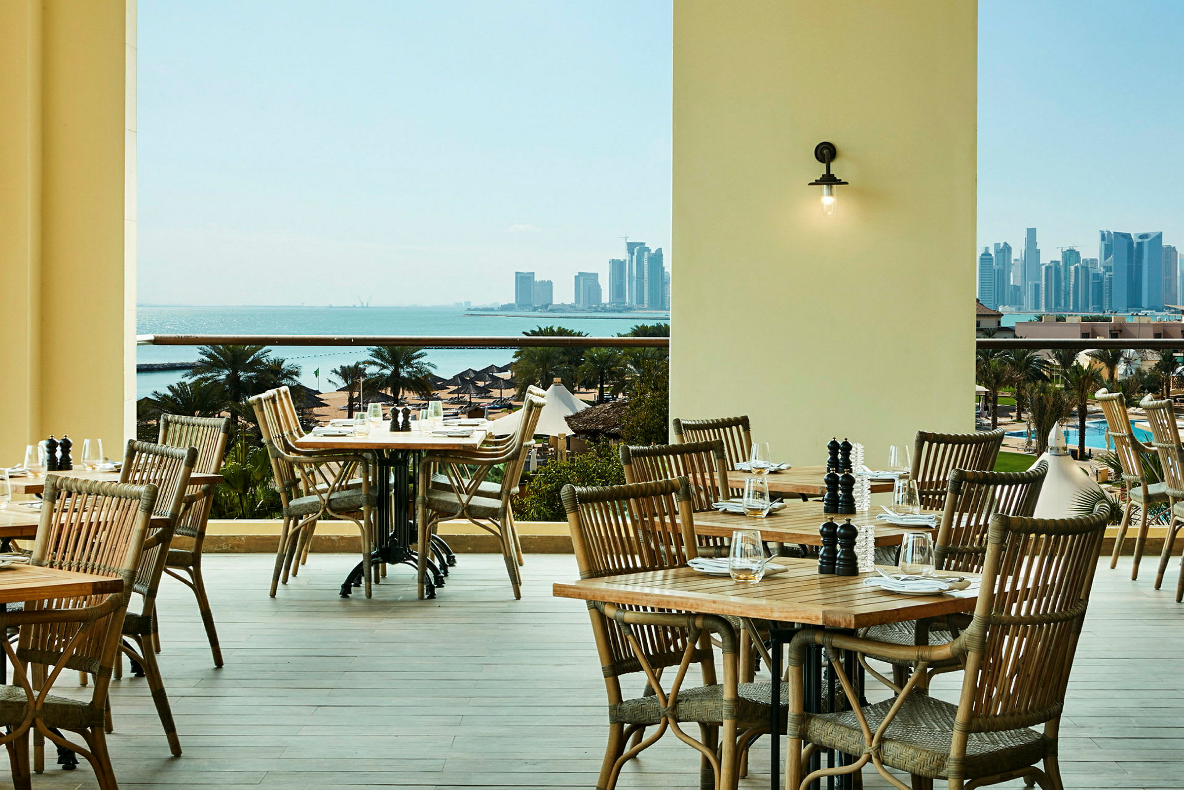 En restaurantterrasse med træmøbler med udsigt over havet og en bysilhuet. (AI-genereret tekst)