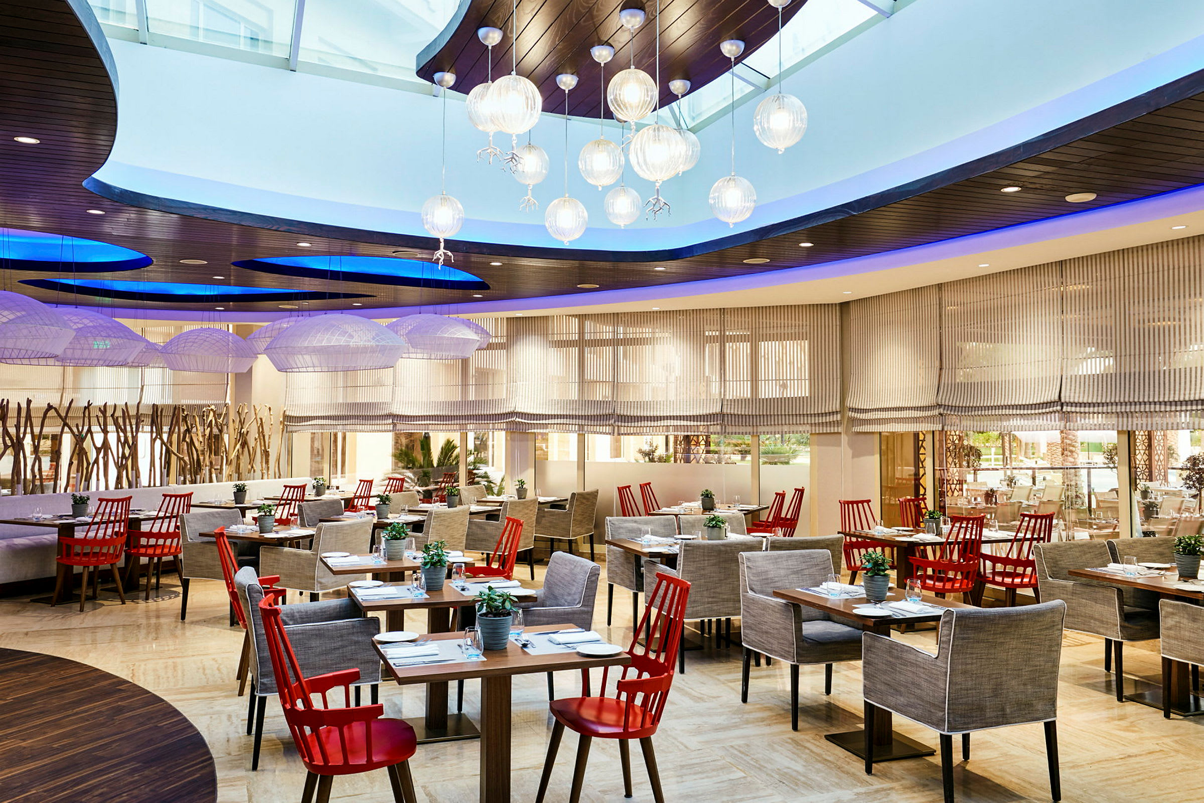 Restaurant med elegant interiør, røde stole og grå lænestole ved flere borde, med moderne lamper i loftet og store vinduer. (AI-genereret tekst)