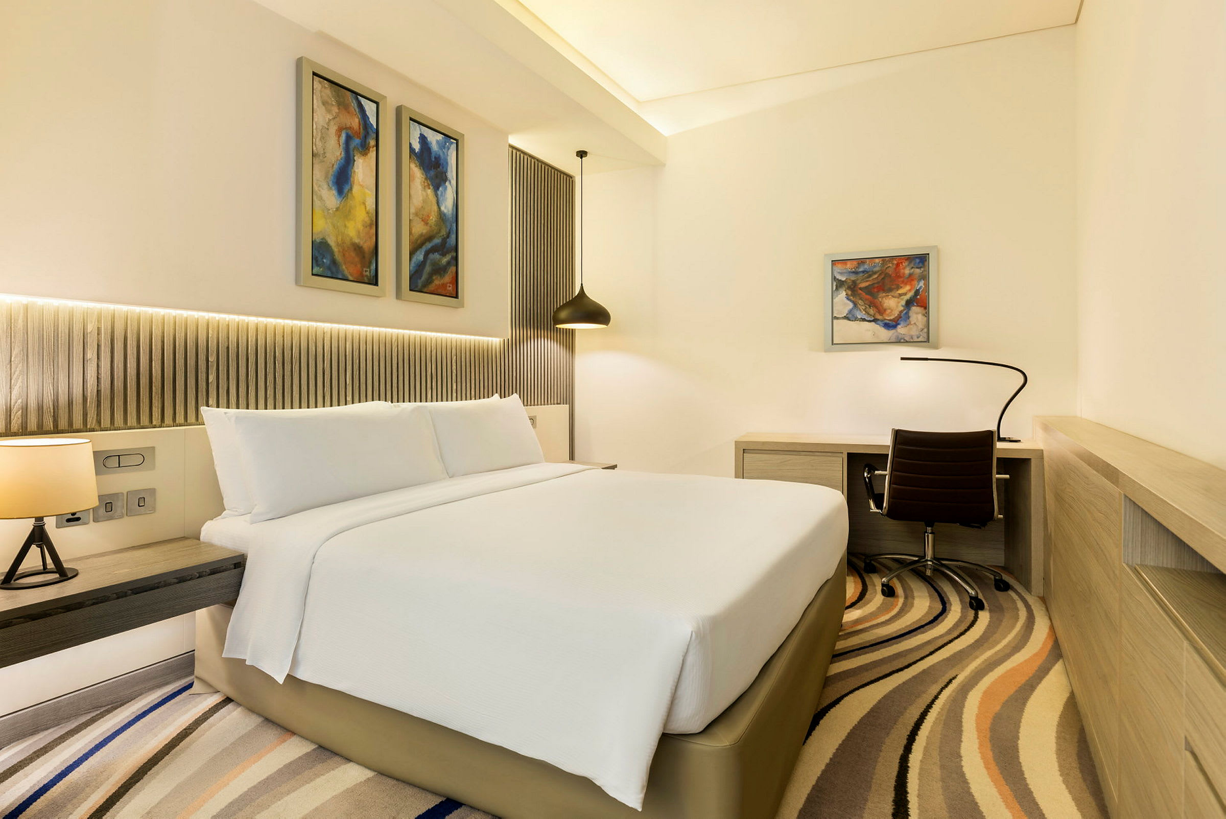 Et moderne hotelværelse med en stor seng, skrivebord og stol samt abstrakt kunst på væggene. (AI-genereret tekst)
