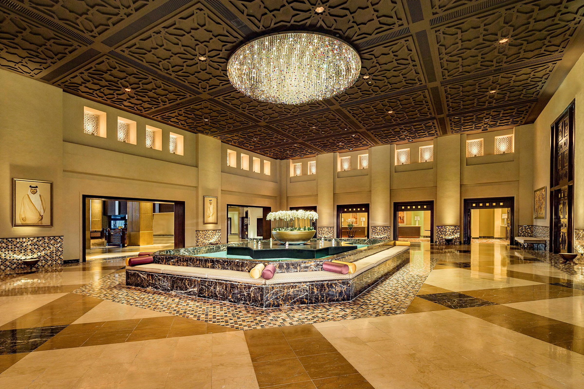 En elegant hotellobby med en stor lysekrone og et centralt springvandsområde omgivet af siddepladser. (AI-genereret tekst)