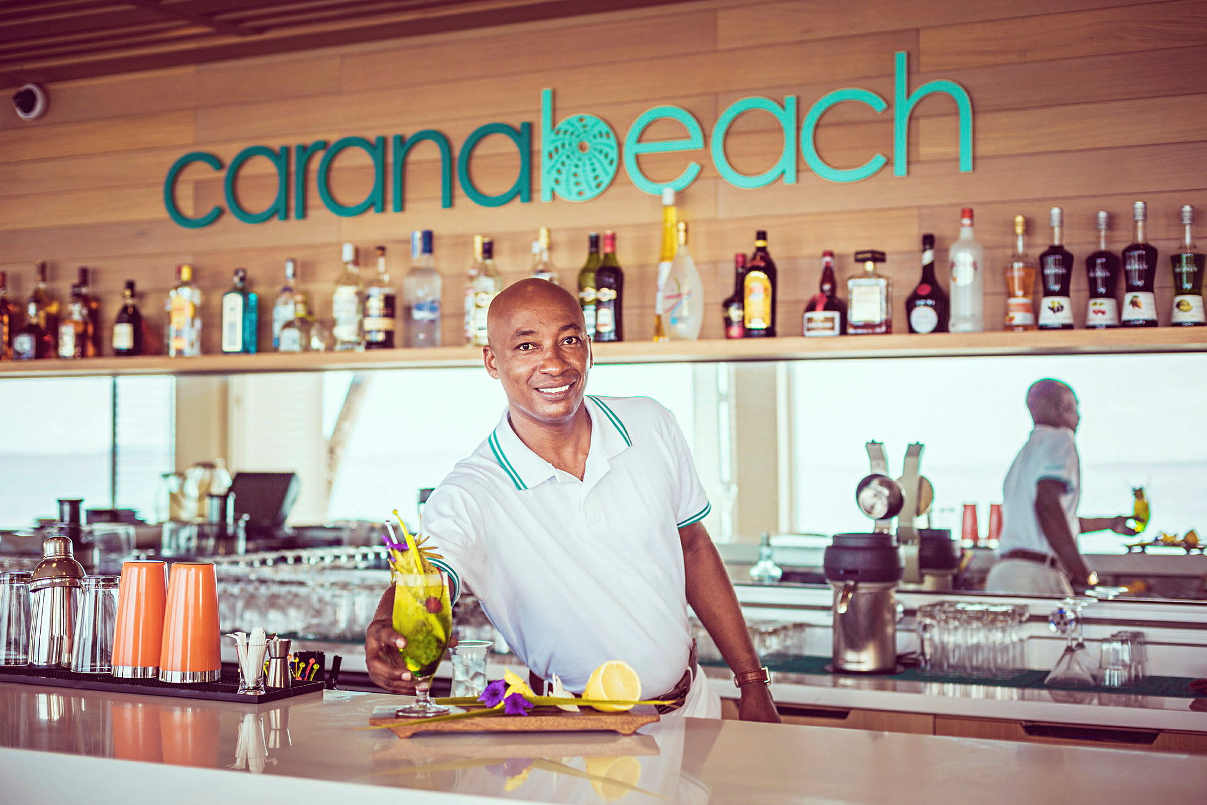 En bartender præsenterer en cocktail på en bar med skiltet 'Carana Beach'. (AI-genereret tekst)