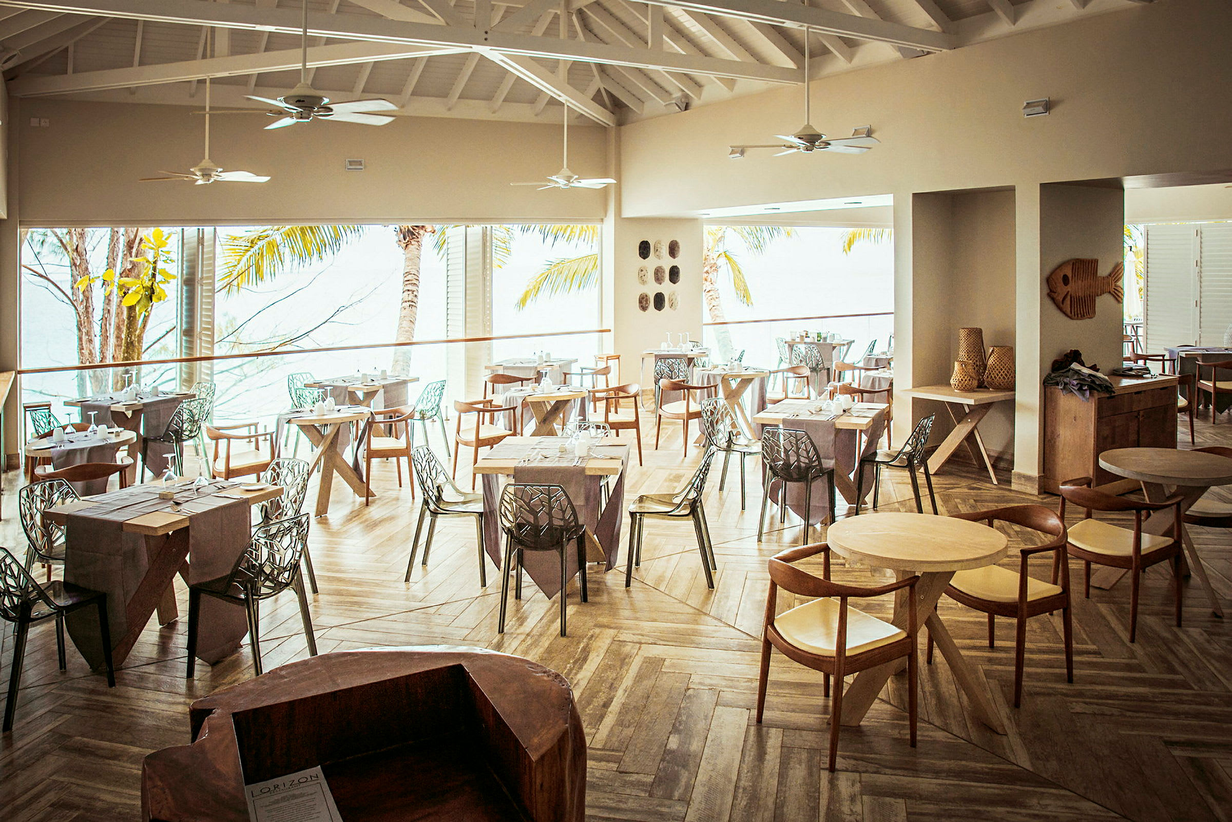 Interiør af en restaurant med træborde og stole samt store vinduer med udsigt til palmer og hav. (AI-genereret tekst)