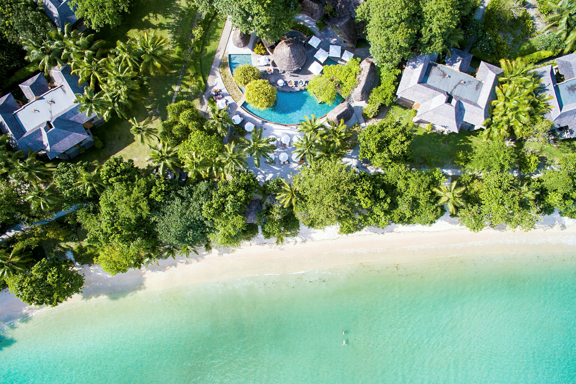 Luftfoto af et resort med en pool, bygninger omgivet af frodig vegetation og en strand ved turkisvand. (AI-genereret tekst)