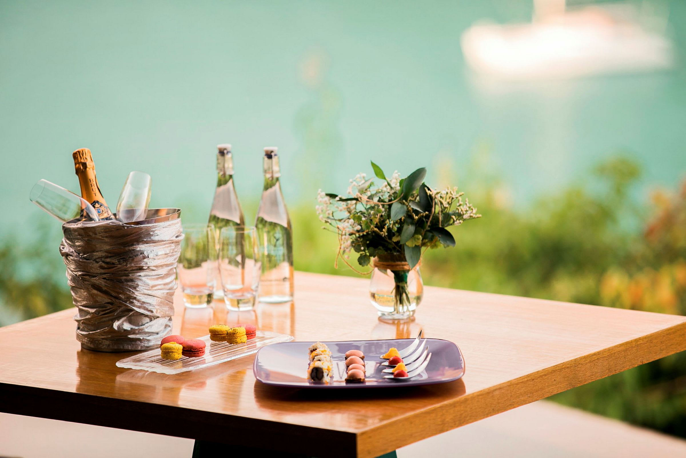 Et træbord med en champagnekøler og glas, vandflasker, en tallerken med mad og småkager samt en vase med blomster, med en sløret baggrund af vand. (AI-genereret tekst)