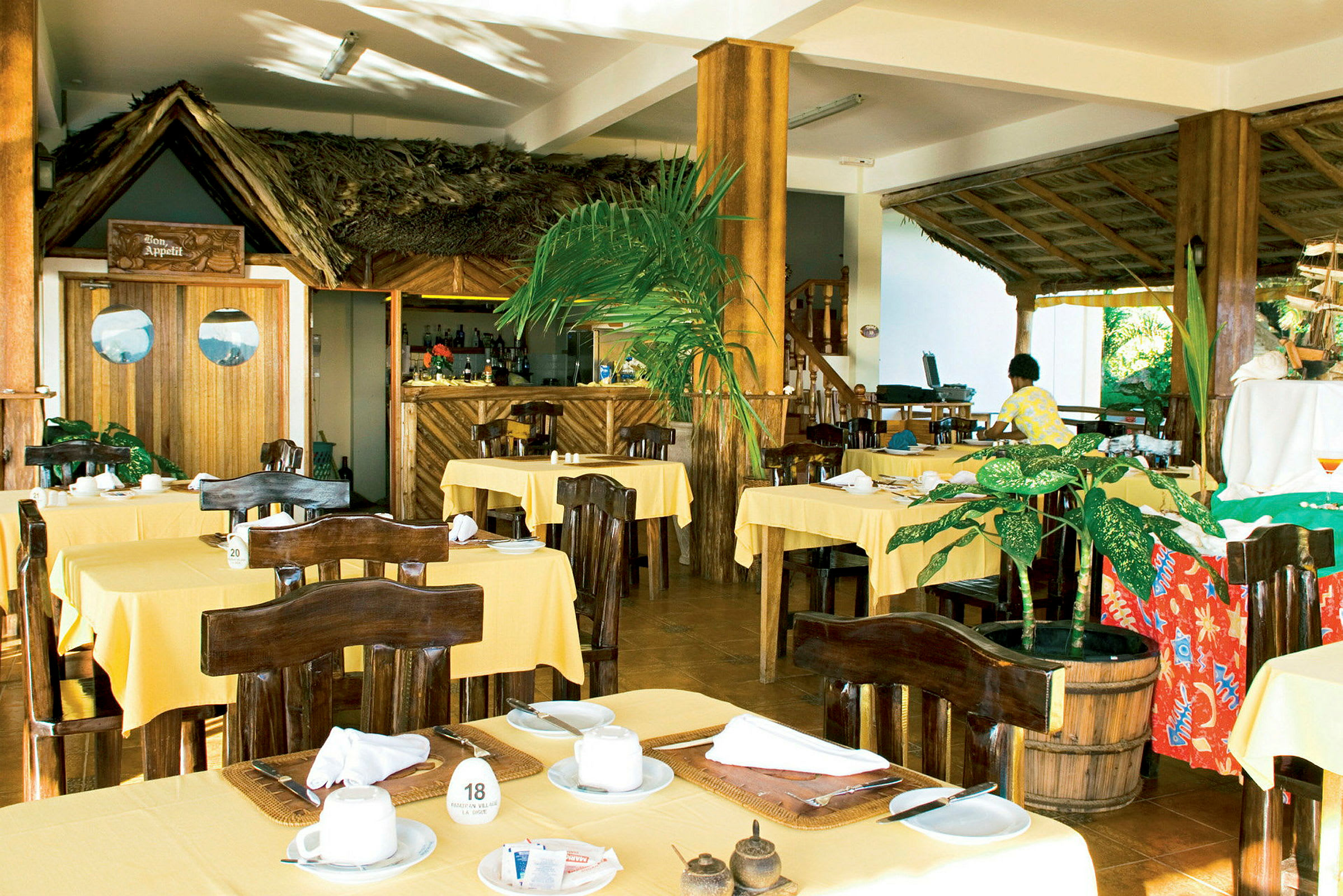 En restaurant med gule duge på bordene og mørkebrune træstole, en bar i baggrunden og dekorationer af tropiske planter. (AI-genereret tekst)