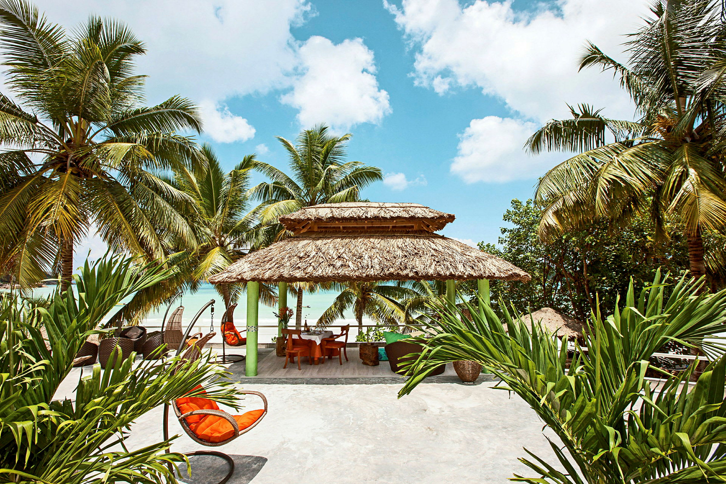 En strandnær hytte med palmebladstag og omgivende tropiske planter. (AI-genereret tekst)