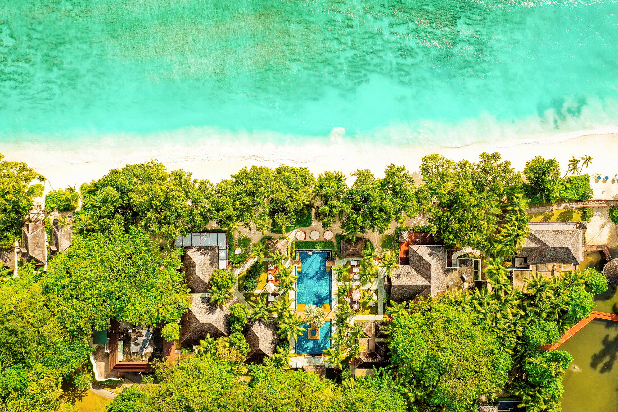 Luftfoto af et hotel ved en strand med pool, omgivet af frodig vegetation. (AI-genereret tekst)