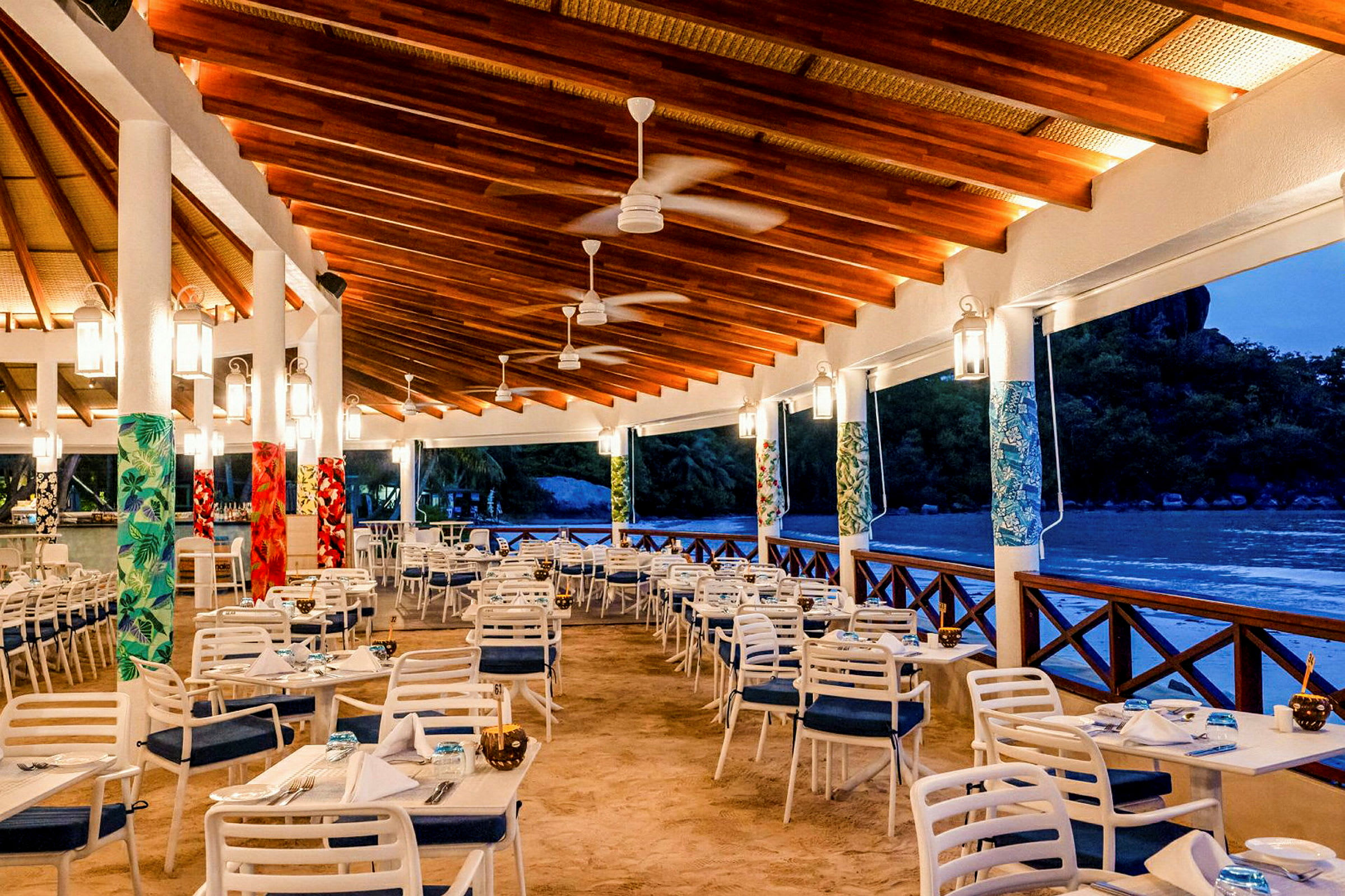 En udendørsrestaurant under tag med borde og stole på sanden, omgivet af tropisk vegetation og med udsigt over en strand. (AI-genereret tekst)