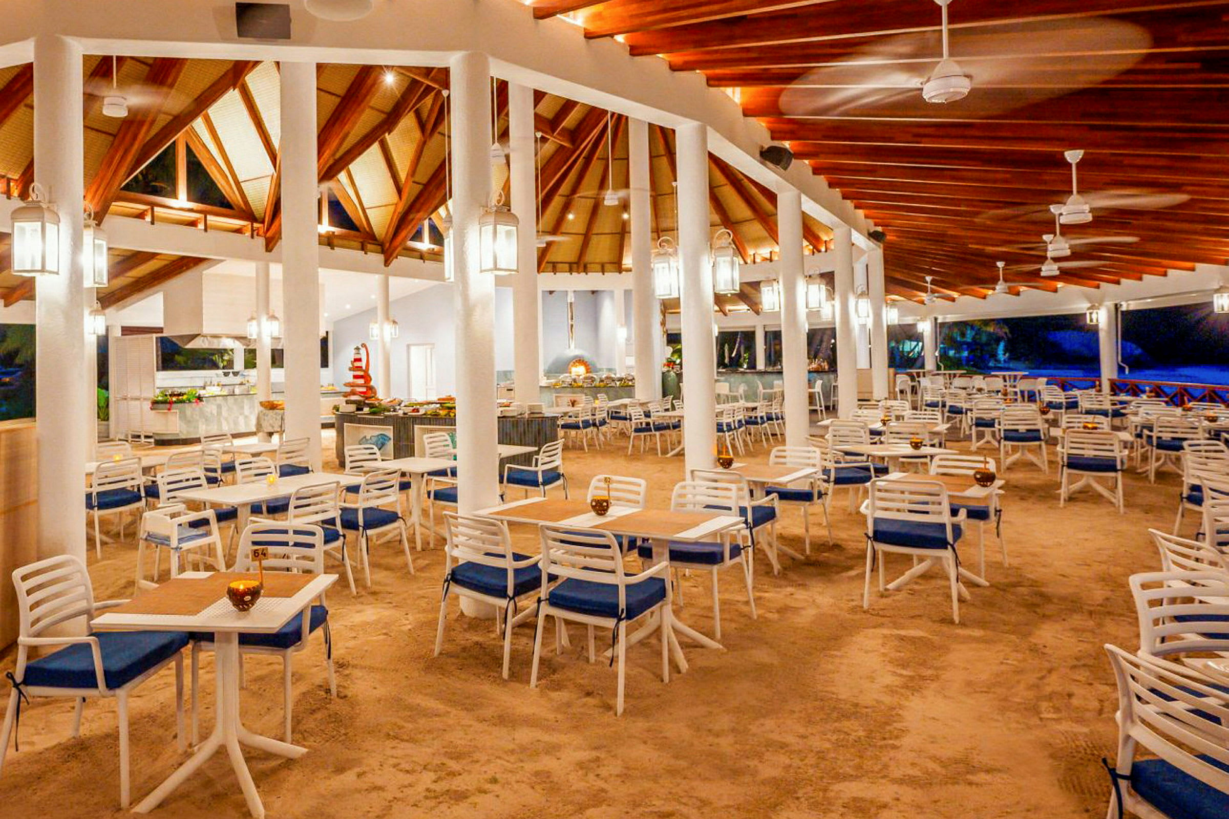 Restaurant med åbent loft af træ, hvide søjler og gulv dækket af sand. Flere borde med blå og hvide stole er arrangeret. (AI-genereret tekst)