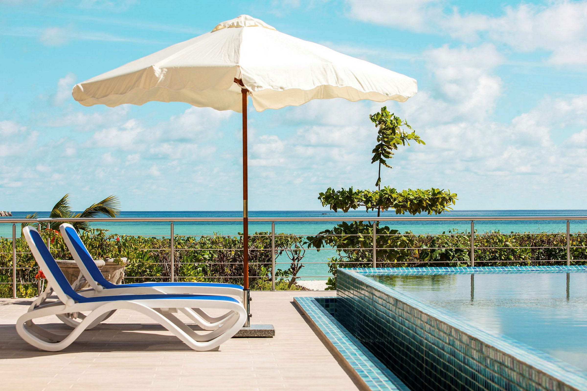 En solrig terrasse med to blå- og hvidstribede liggestole, en parasol og en pool, med udsigt over havet. (AI-genereret tekst)