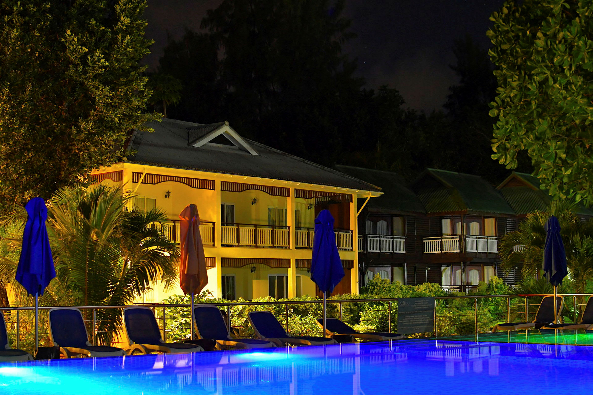En natteudsigt med en oplyst bygning og en swimmingpool med solsenge og parasoller. (AI-genereret tekst)