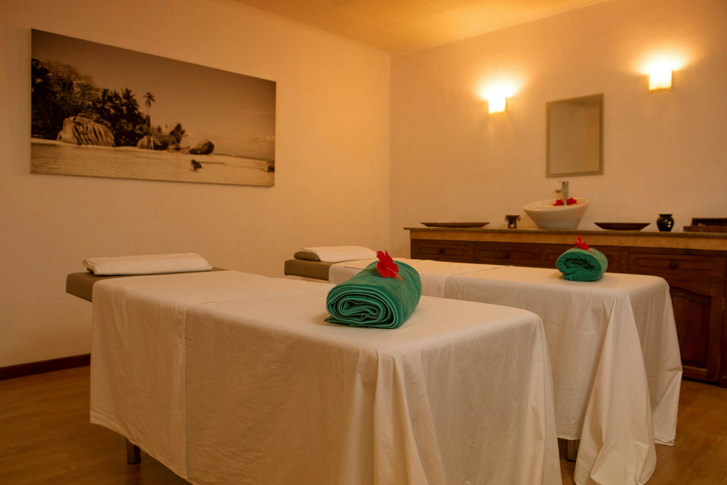 Interiør af et spa med to massageborde dækket med hvide duge og grønne håndklæder. I baggrunden ses et bord med en skål. (AI-genereret tekst)