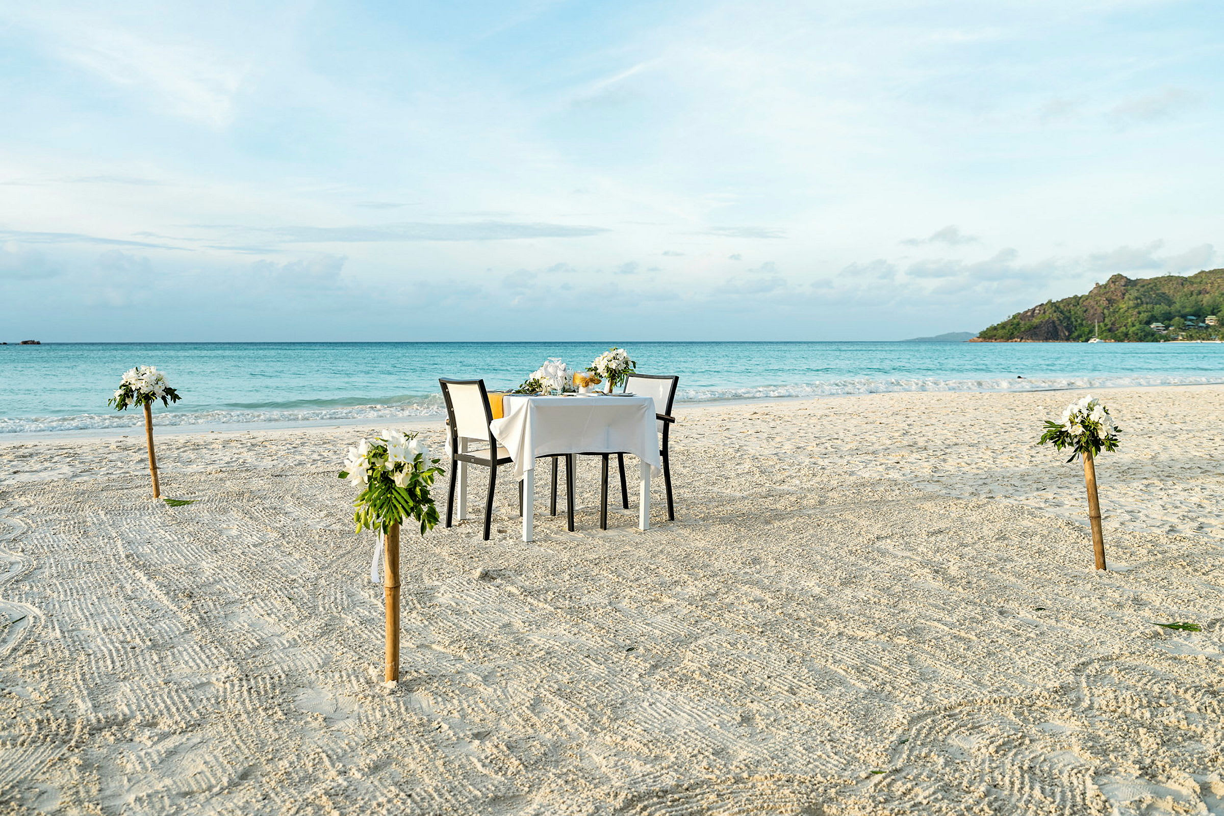 Et dækket bord med to stole står på en sandstrand. Bordet er dekoreret med blomsterdekorationer, og fire blomsterdekorationer er placeret på stolper rundt om bordet. I baggrunden ses havet og en grøn klippe. (AI-genereret tekst)