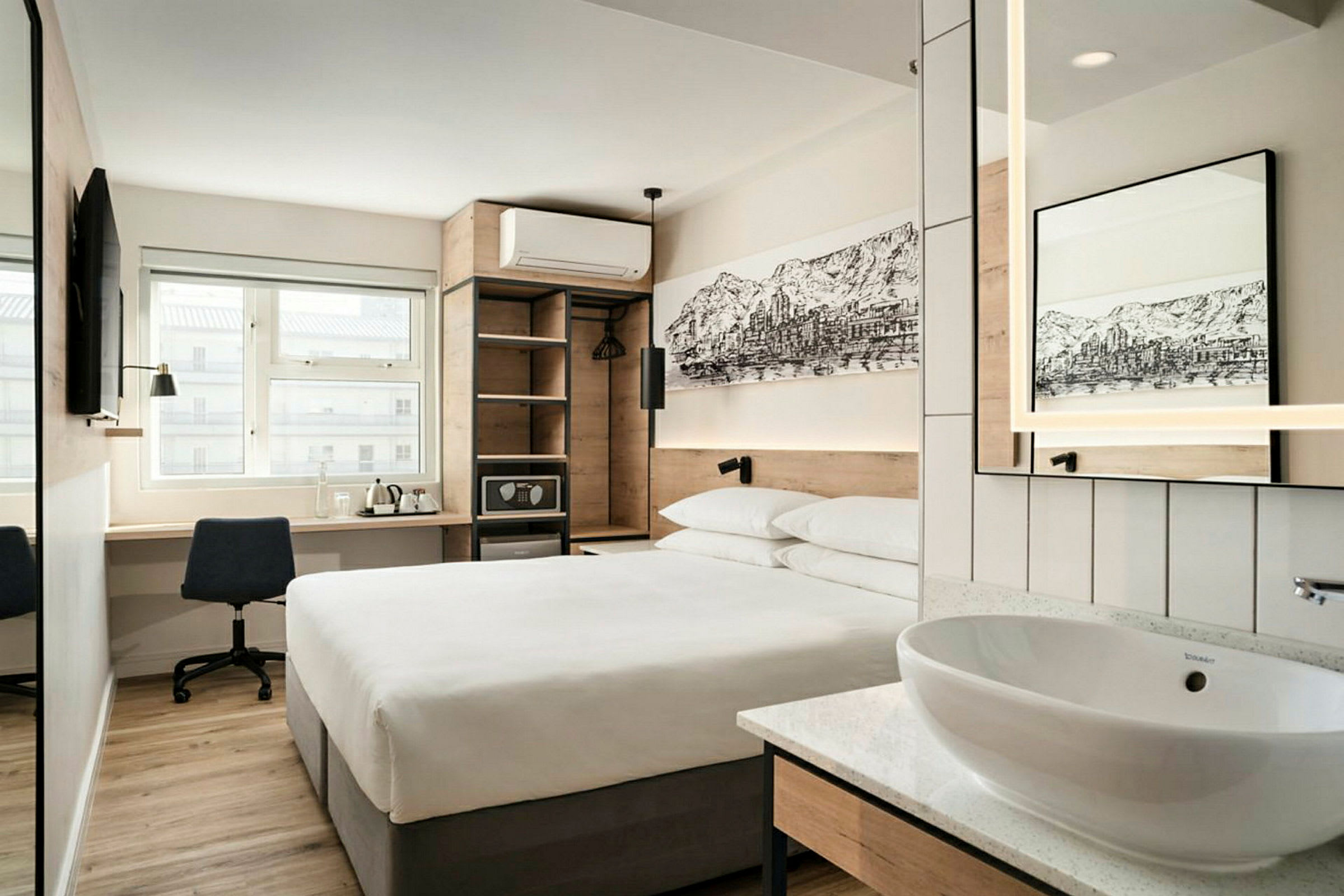 Et moderne hotelværelse med en dobbeltseng, et skrivebord med stol, et vægmonteret TV, samt en åben garderobe og en lille håndvask med spejl. (AI-genereret tekst)