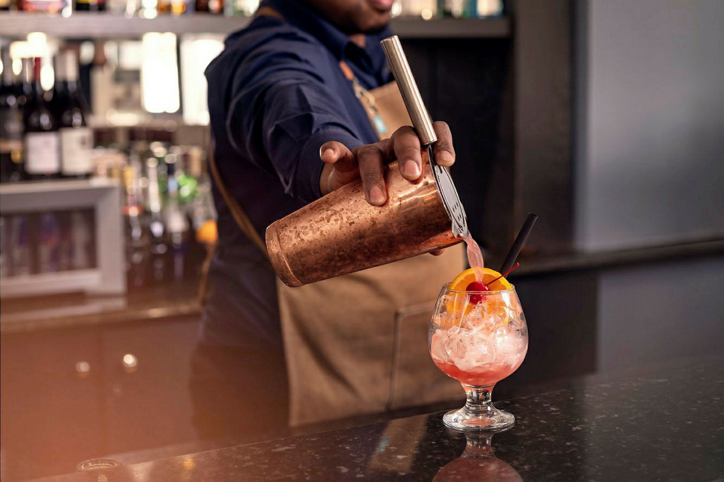 En bartender hælder en lyserød drik fra en shaker i et glas med is og en dekoration af appelsinskive og kirsebær. (AI-genereret tekst)