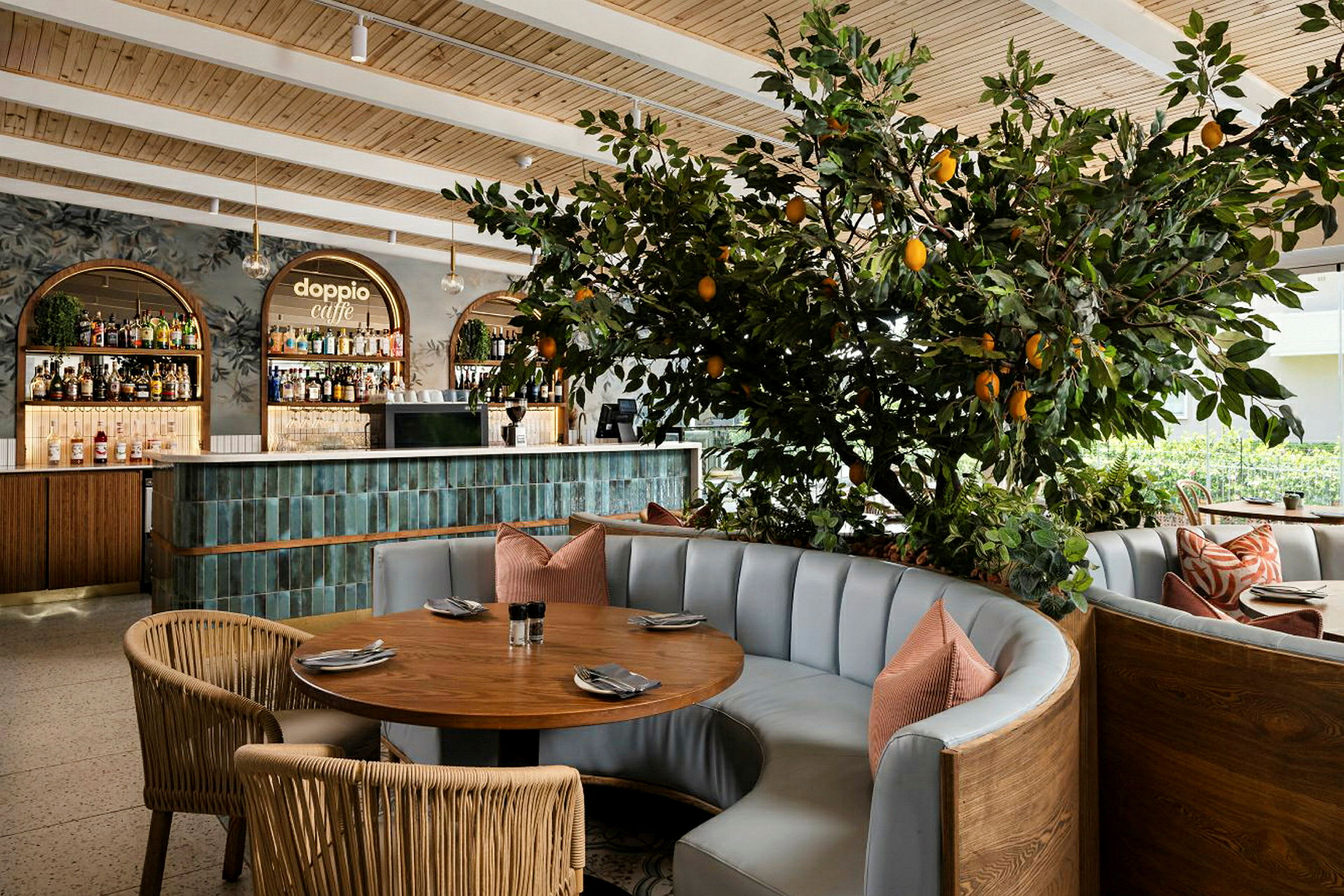 Interiør i en restaurant med et rundt bord, polstrede sofaer og en dekorativt plantet citrusplante med frugter. I baggrunden ses en bardisk. (AI-genereret tekst)