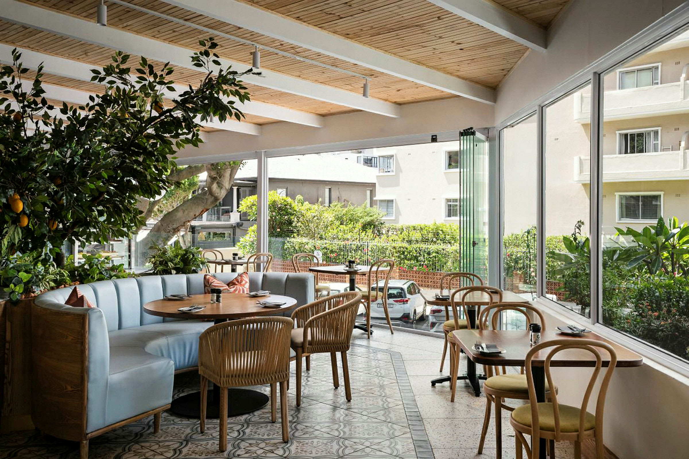 En café eller restaurant med store vinduer, der lader masser af lys komme ind. Lokalet har trædetaljer i loftet, bøjet træstole og et langbord med polstrede siddepladser. (AI-genereret tekst)