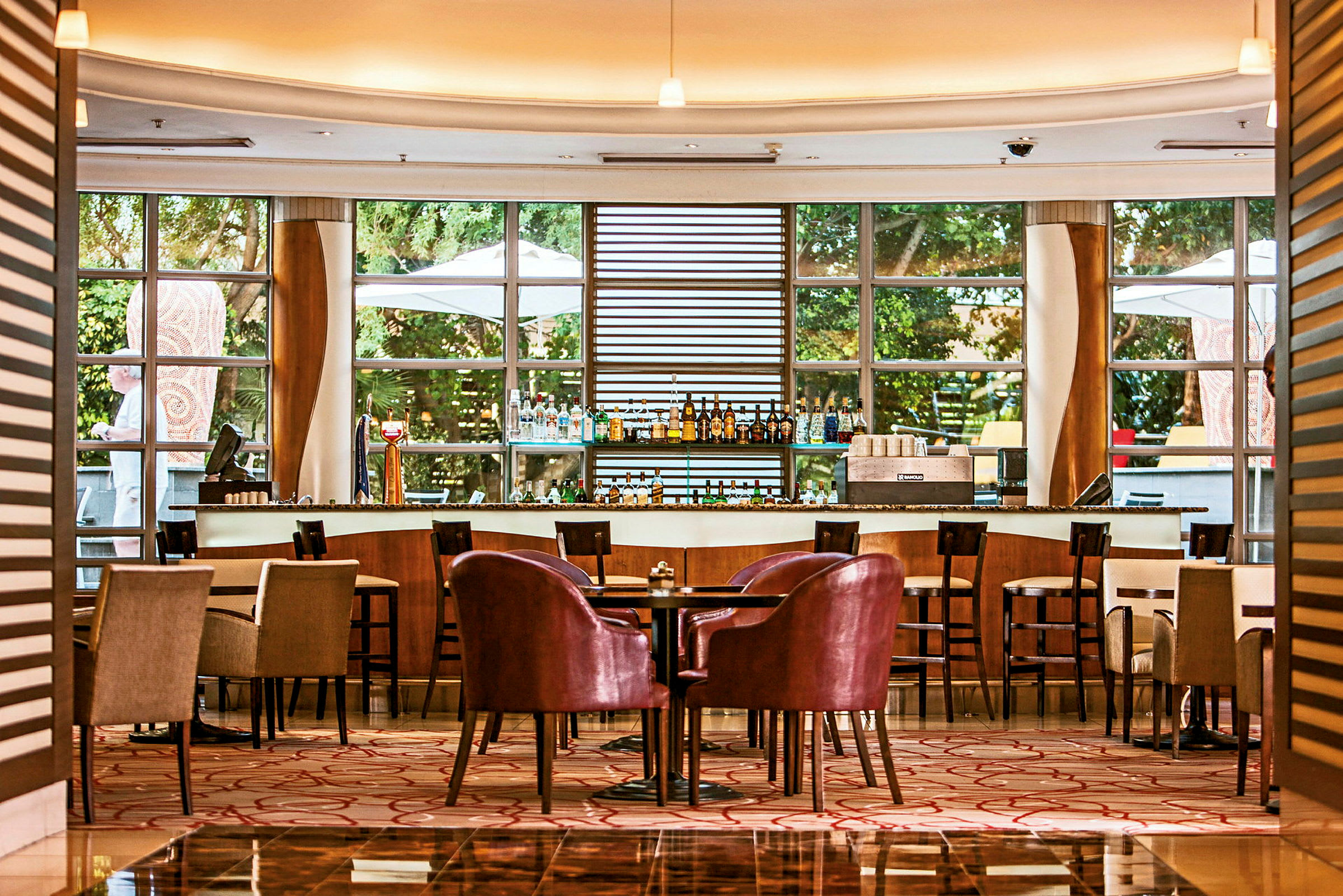 En elegant bar og siddeområde med borde og stole på et hotel eller en restaurant. Store vinduer lukker masser af lys ind udefra. (AI-genereret tekst)
